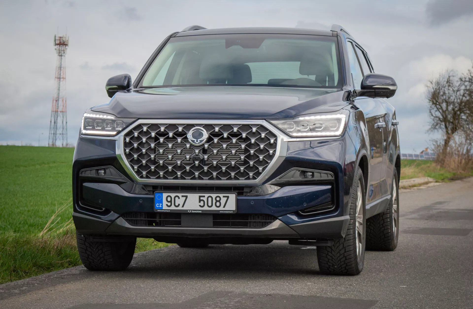 SUV SsangYong Rexton 2021 sur une route de campagne, présentant une vue détaillée de son design avant avec une grille géométrique et des phares LED, reflétant une technologie avancée et des capacités tout-terrain