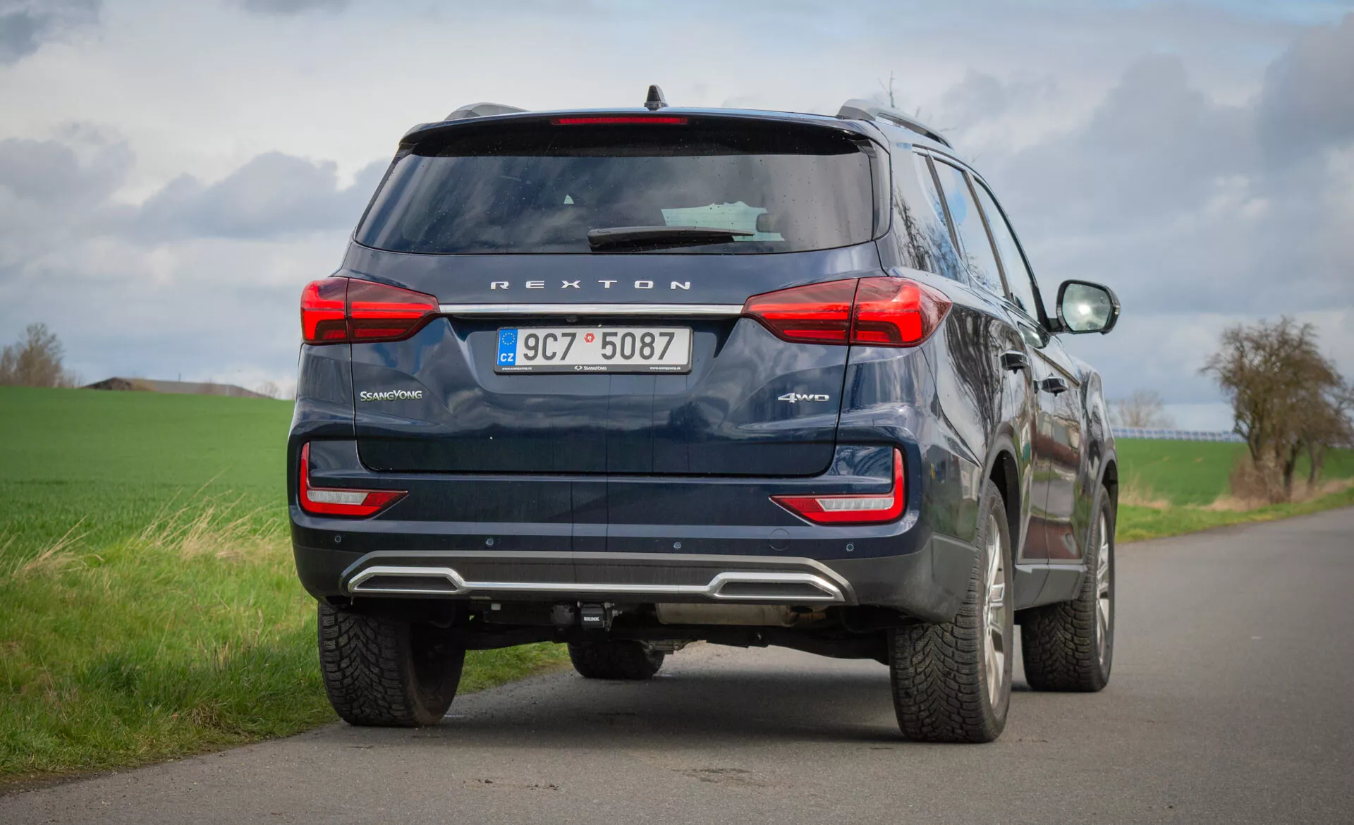 Vue arrière d'un SUV bleu SsangYong Rexton 2021 garé sur une route rurale à deux voies, mettant en évidence ses caractéristiques de conception telles que les feux arrière, la fenêtre arrière, les tuyaux d'échappement et la technologie de sécurité, y compris la caméra de recul et les capteurs de stationnement, sous un ciel nuageux.