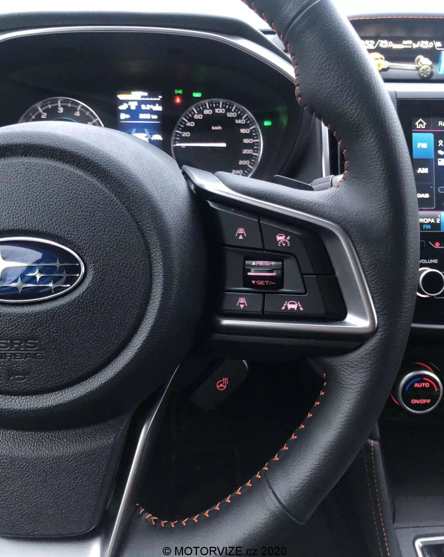 Vue intérieure d'une Subaru XV 2018 depuis le siège du conducteur, mettant en évidence le volant en cuir noir avec des coutures orange, doté de boutons de commande multimédia et d'un tableau de bord partiellement visible avec des affichages analogiques et numériques. La console centrale avec son écran tactile d'infodivertissement et le contrôle automatique de la climatisation bi-zone est également visible en arrière-plan.