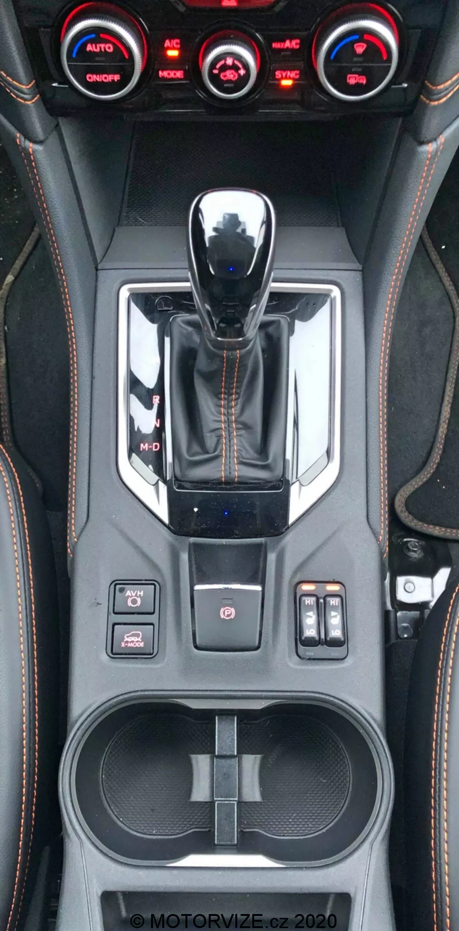 Vue rapprochée verticale de la console centrale de la Subaru XV 2018, montrant le levier de vitesses avec finition métallique, le sélecteur de mode de conduite, divers boutons de commande, y compris le contrôle de traction, les capteurs de stationnement et les réglages de suspension, un système de climatisation à trois zones, des détails cousus à la main en similicuir, un porte-gobelets double, un espace de rangement avec ports USB et un filigrane visible MOTORVIZE.cz.