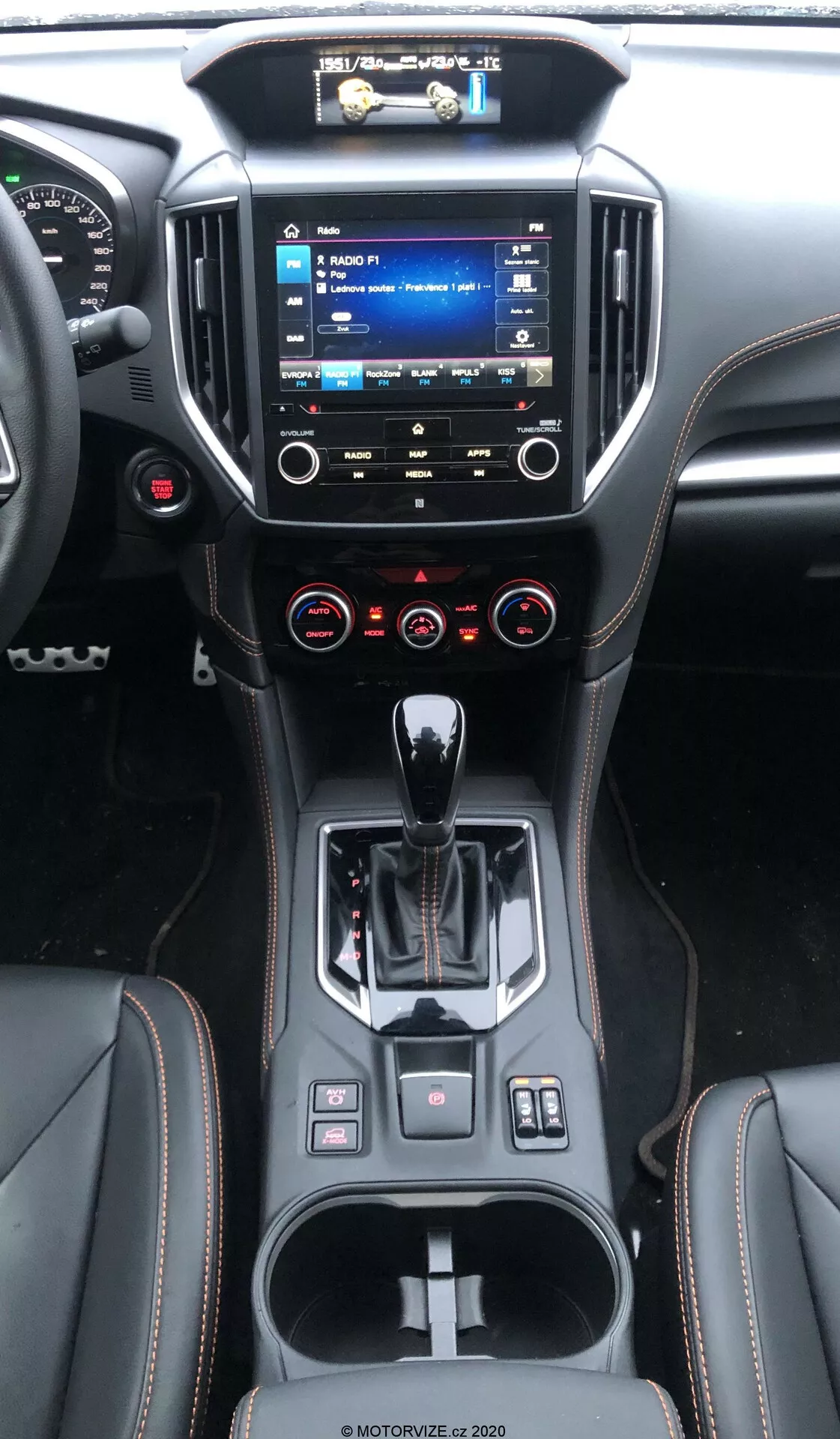 L'intérieur d'une Subaru XV 2018 (subaru-xv-1-6i-trend-lineartronic) montrant la console centrale et le tableau de bord depuis la perspective du siège arrière, avec un système d'infodivertissement surélevé et des menus visibles, un contrôle climatique à double zone, un sélecteur de vitesse automatique ergonomique, un design contemporain des matériaux, des porte-gobelets et de petits espaces de rangement, divers boutons de contrôle et un volant multifonctions.