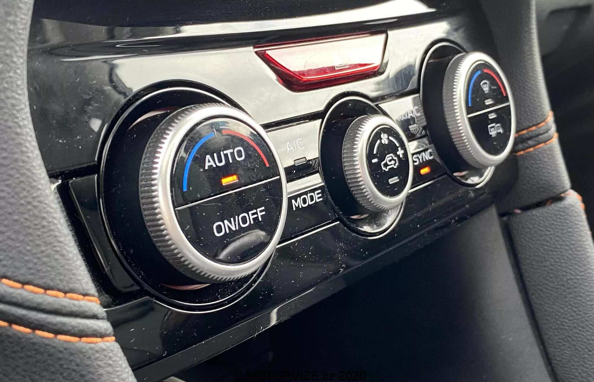Gros plan des boutons de commande de la climatisation dans l'intérieur de la Subaru XV 2018. Pris de la perspective du conducteur, il montre trois boutons circulaires : celui de gauche en mode 'AUTO' pour la vitesse du ventilateur, celui du centre pour la sélection du mode 'ON/OFF' et celui de droite pour le contrôle de la température du passager en mode 'SYNC'. Au-dessus du bouton central se trouve un bouton de détresse rouge. L'image met en avant des accents chromés sur les boutons et des surpiqûres rouges en dessous.