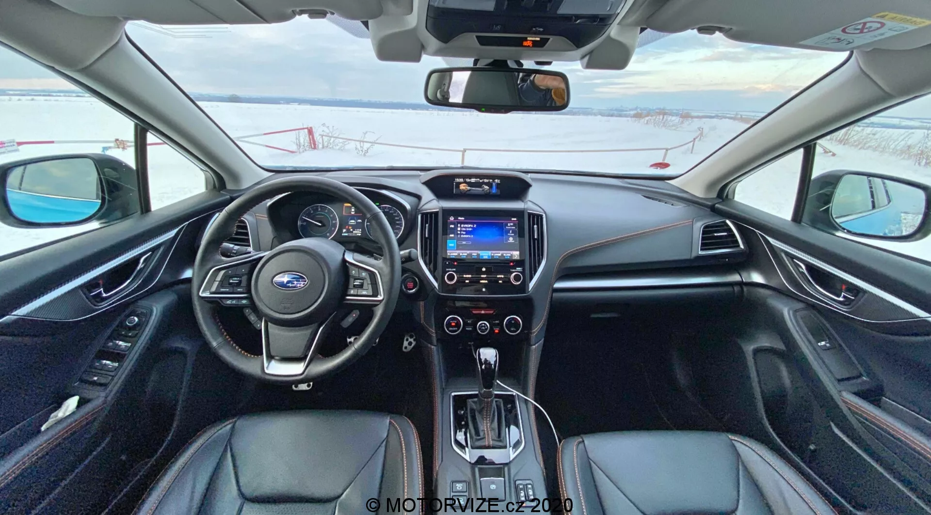 Vue intérieure d'une Subaru XV 2018 (Subaru XV 1.6i Trend Lineartronic) depuis le siège du conducteur montrant le volant en cuir avec commandes intégrées, tableau de bord analogique et numérique partiellement visible, écran central d'info-divertissement affichant une interface bleue, boutons de commande du climat, levier de vitesses automatique entouré de boutons opérationnels, sellerie noire avec accents argentés, commandes de chauffage des sièges et rétroviseur avec écran numérique. La voiture est stationnée et il y a de la neige à l'extérieur, indiquant un climat froid.