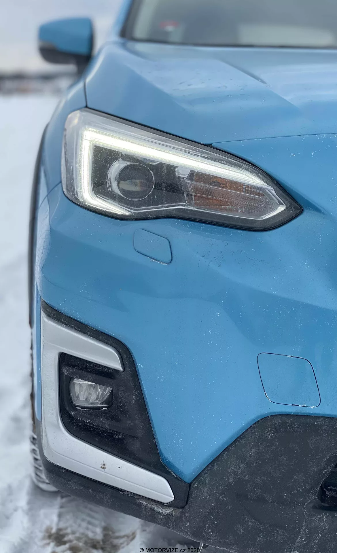 Vue en gros plan du côté avant gauche d'une Subaru XV 1.6i Trend Lineartronic bleue de 2018, prise sous un angle oblique se concentrant sur le phare et une partie de la calandre et du pare-chocs avant. Le phare avant gauche proéminent dispose d'une lentille de projecteur indicatrice de systèmes HID ou LED et de feux de jour à LED intégrés. L'image montre également une partie du pare-chocs avant qui abrite probablement un éclairage supplémentaire. La carrosserie du véhicule présente des lignes nettes et des formes aérodynamiques, sur un fond hivernal avec une légère couche de neige.