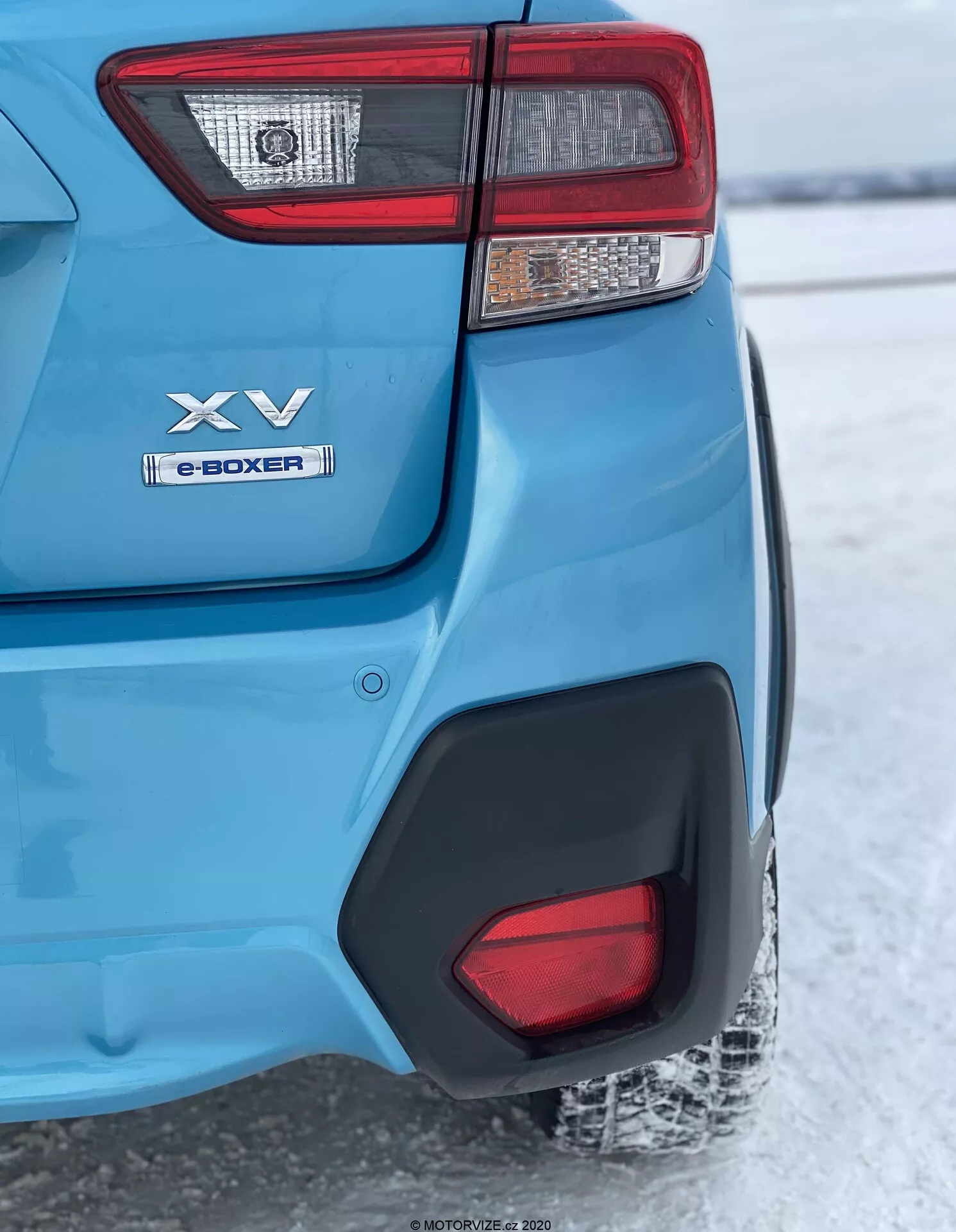 Un gros plan en angle arrière du modèle Subaru XV 2018 bleu, se concentrant sur le coin arrière gauche de la lumière arrière à la roue arrière. Les caractéristiques clés incluent un ensemble de feux arrière complexe, des badges 'XV' et 'e-BOXER' indiquant la technologie hybride, une peinture bleue métallisée brillante avec un moulage d'arche de roue en plastique noir, un insert en plastique noir sur le pare-chocs arrière reflétant un sol enneigé suggérant un climat froid, et un pneu arrière partiellement visible avec une bande de roulement recouverte de neige. La mise au point est nette sur le coin arrière tandis que l'arrière-plan est légèrement flou en raison de la profondeur de champ.