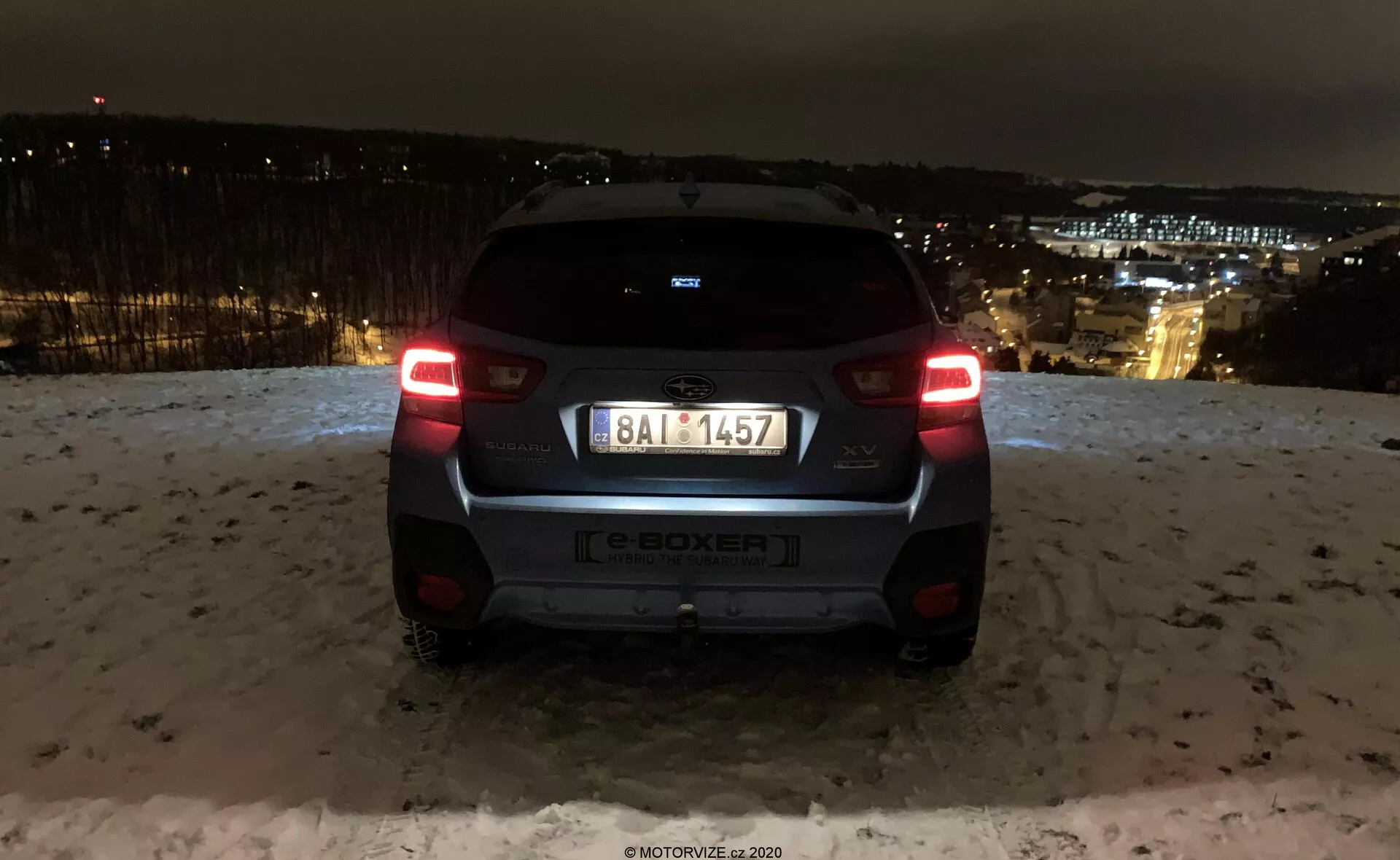 Vue arrière de la Subaru XV 2018 (subaru-xv-1-6i-trend-lineartronic) capturée de nuit avec les lumières de la ville en arrière-plan. La voiture est située sur une zone surélevée, montrant les feux arrière illuminés, la lunette arrière, le badge du fabricant, l'indication du modèle e-BOXER, la plaque d'immatriculation, le pare-chocs arrière et la caméra ou le capteur de stationnement. Les surfaces réfléchissantes et la neige au sol suggèrent des conditions hivernales. Le design moderne du véhicule comprend un petit spoiler, un essuie-glace arrière minimaliste et un pare-chocs bicolore, mettant en valeur les éléments d'éclairage et de design de la marque automobile.