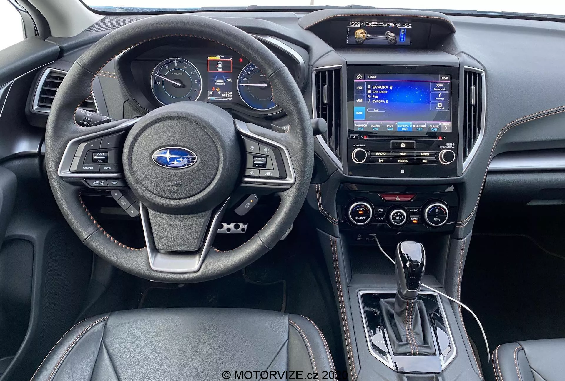 Vue intérieure depuis le siège arrière d'une Subaru XV 1.6i Trend Lineartronic 2018. Comprend un volant moderne avec boutons de commande audio et de régulateur de vitesse, un tableau de bord partiellement visible, un système d'infodivertissement à écran tactile avec navigation, des commandes de climatisation à double zone, un levier de vitesses automatique en position de parking, un interrupteur de frein de parking électronique, des commandes de chauffage des sièges et un intérieur en cuir noir bicolore avec des accents argentés.