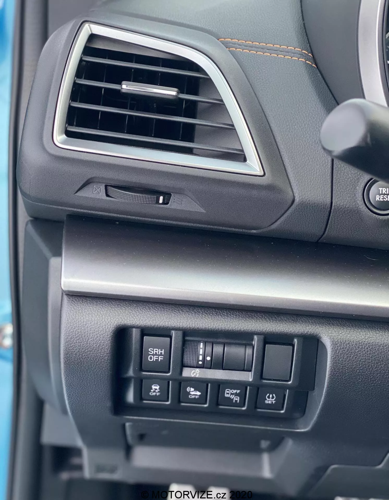 Vue rapprochée de la planche de bord côté conducteur de la Subaru XV 1.6i Trend Lineartronic 2018. L'image se concentre sur une bouche d'aération rectangulaire aux bords arrondis avec un ajusteur directionnel, un volant partiellement visible avec des boutons de commande audio, et un compartiment fermé avec une surface doucement texturisée sous la bouche d'aération. Les boutons de commande pour des fonctions telles que les phares adaptatifs (bouton SRH OFF) et d'autres réglages du véhicule sont également visibles. Les matériaux de la planche de bord semblent être en plastique noir ou gris foncé avec différentes textures, indiquant durabilité et qualité.