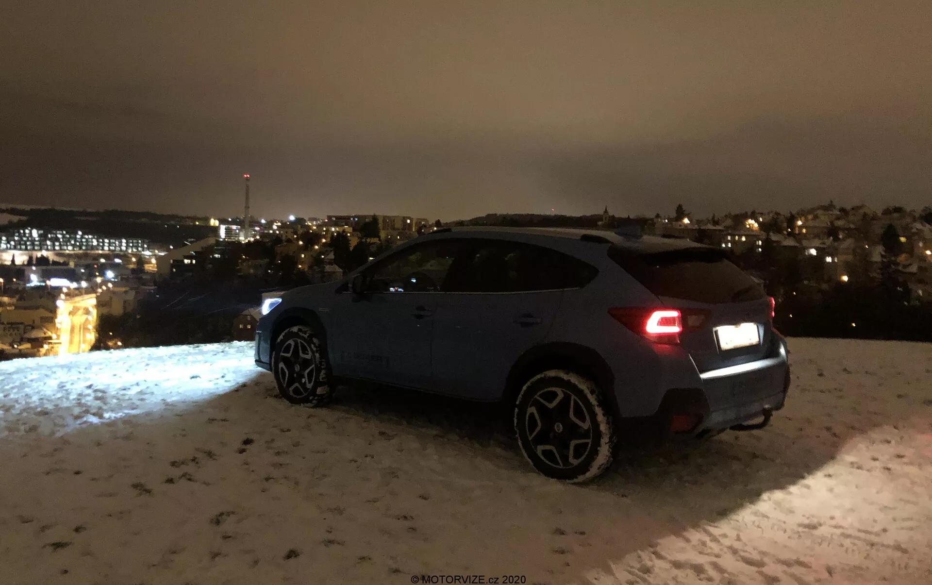 Subaru XV 1.6i Trend Lineartronic 2018 photographié de nuit sous un angle de vue arrière trois-quarts, montrant le côté droit et l'arrière, y compris les feux arrière, le pare-chocs arrière, la roue arrière droite, les portes passager et la roue avant droite. Le véhicule, probablement de couleur bleu foncé ou gris, est garé sur une surface couverte de neige avec des lumières de la ville visibles au loin. Les feux arrière suggèrent que la voiture pourrait être allumée, et les roues distinctives plus grandes indiquent un niveau de finition supérieur ou une variante sportive. La scène met l'accent sur les composants esthétiques et l'ambiance du cadre sans visibilité claire des fonctionnalités technologiques avancées.