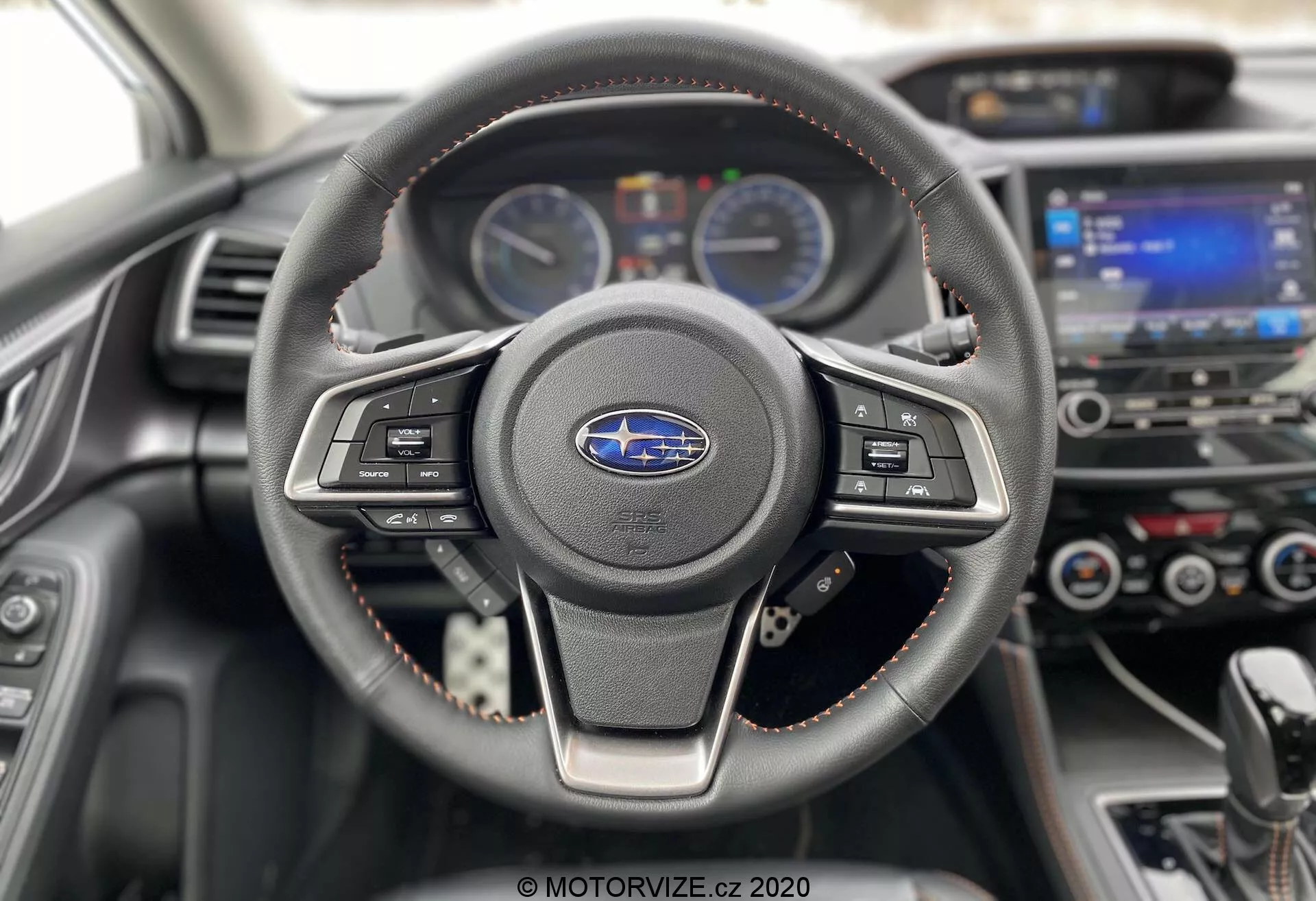 Vue intérieure d'une Subaru XV 2018 (Subaru-xv-1-6i-trend-lineartronic) centrée sur le volant et le tableau de bord. Le volant à trois branches, recouvert de cuir avec des coutures visibles, comporte des boutons de commande multimédia et de régulateur de vitesse. Derrière, le tableau de bord affiche deux jauges analogiques séparées par un écran numérique d'information conducteur. En arrière-plan, légèrement flou, se trouvent le système d'infodivertissement à écran tactile et les commandes de climatisation. Les coutures rouges sur le volant et les garnitures du tableau de bord mettent en avant un niveau de finition sport ou premium. Prise depuis le siège conducteur, cette image moderne et bien entretenue met l'accent sur la technologie et le design automobile.