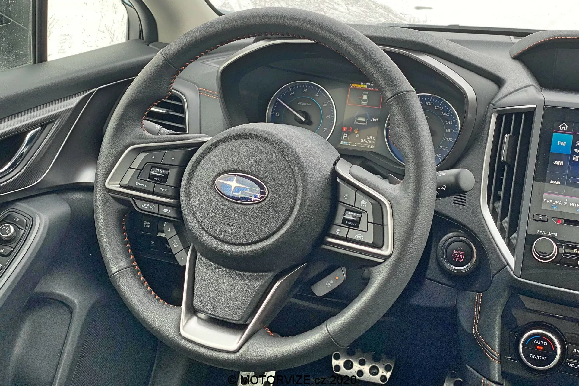 Vue intérieure du Subaru XV 2018, concentrée sur le volant et la zone du tableau de bord. Volant central à trois branches avec commandes intégrées, tableau de bord numérique affichant le compteur de vitesse, le compte-tours et les écrans d'information. Console centrale avec système d'infotainment affichant une carte et commandes de climatisation en dessous. Accents métalliques, volant en cuir, détails rouges autour des aérateurs. Boutons de commande supplémentaires à gauche, pédales de style sport visibles en bas à gauche. Prise de vue frontale, légèrement vers le bas, montrant les commandes et la technologie du conducteur.