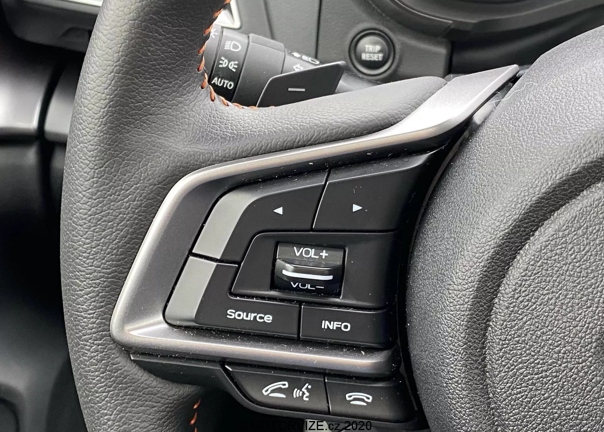 Vue rapprochée des commandes du volant de la Subaru XV 2018 depuis la perspective du siège conducteur. L'image met en évidence les boutons multifonctions sur le rayon gauche, y compris les boutons de réglage du volume, le bouton INFO pour les paramètres de l'écran et une icône ressemblant à un menu. Les interrupteurs du régulateur de vitesse sont légèrement flous au-dessus. Les commandes sont en plastique et caoutchouc, avec un volant en cuir présentant un design ergonomique. Les métadonnées en bas à droite indiquent 'IZE-EV2020'.