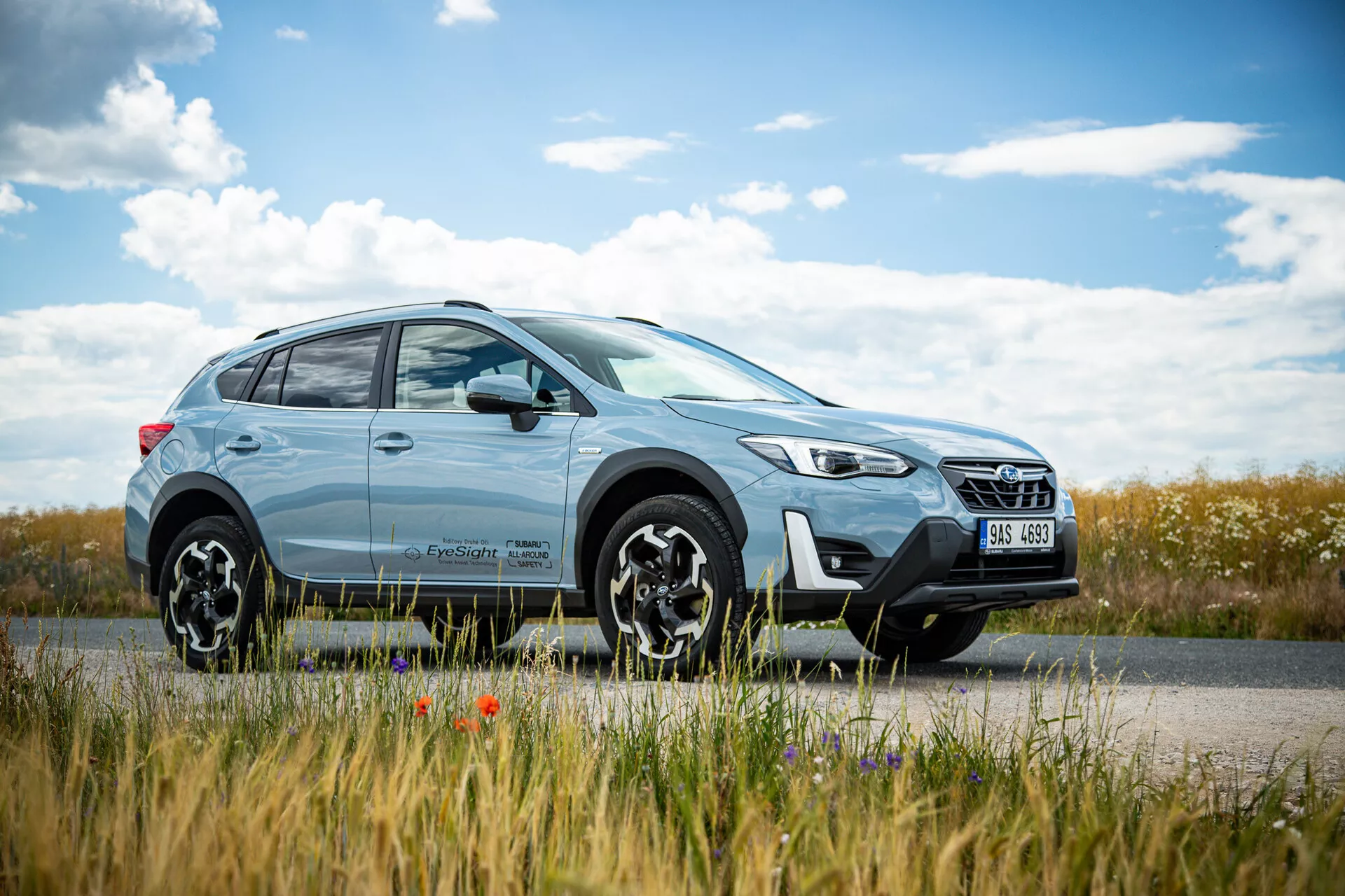 SUV crossover Subaru XV 2021 bleu clair stationné en bord de route avec un paysage champêtre en arrière-plan, présentant un design moderne avec des phares LED et des roues en alliage à multiples branches, soulignant les capacités à la fois urbaines et tout-terrain.