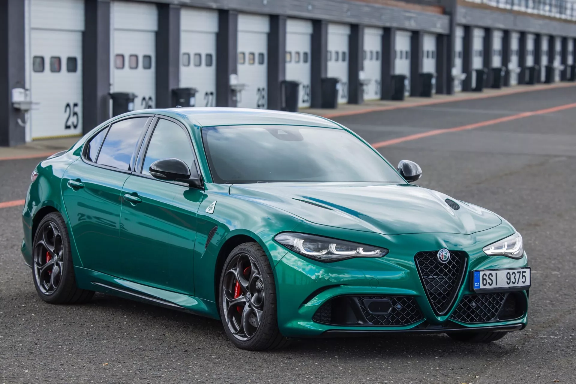 Berline sportive Alfa Romeo Giulia Quadrifoglio 2023 avec finition métallique, vue sous un angle avant de trois quarts dans la voie des stands d'un circuit de course, montrant sa grille avant distinctive, phares LED élégants, grandes entrées d'air, jantes en alliage à plusieurs branches avec étriers de frein rouges, profil aérodynamique sportif, conception avancée de rétroviseurs latéraux et dessin de bande de roulement des pneus orienté performance.