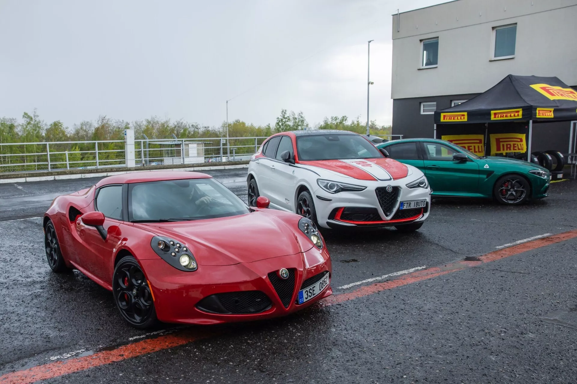 Alfa-Romeo Giulia Quadrifoglio 2023, coupé sportif rouge garé dans la zone des stands d'un circuit sous un ciel nuageux, montrant un profil avant et latéral avec phares LED, grille frontale triangulaire avec logo, grandes entrées d'air sur le pare-choc et jantes en alliage.