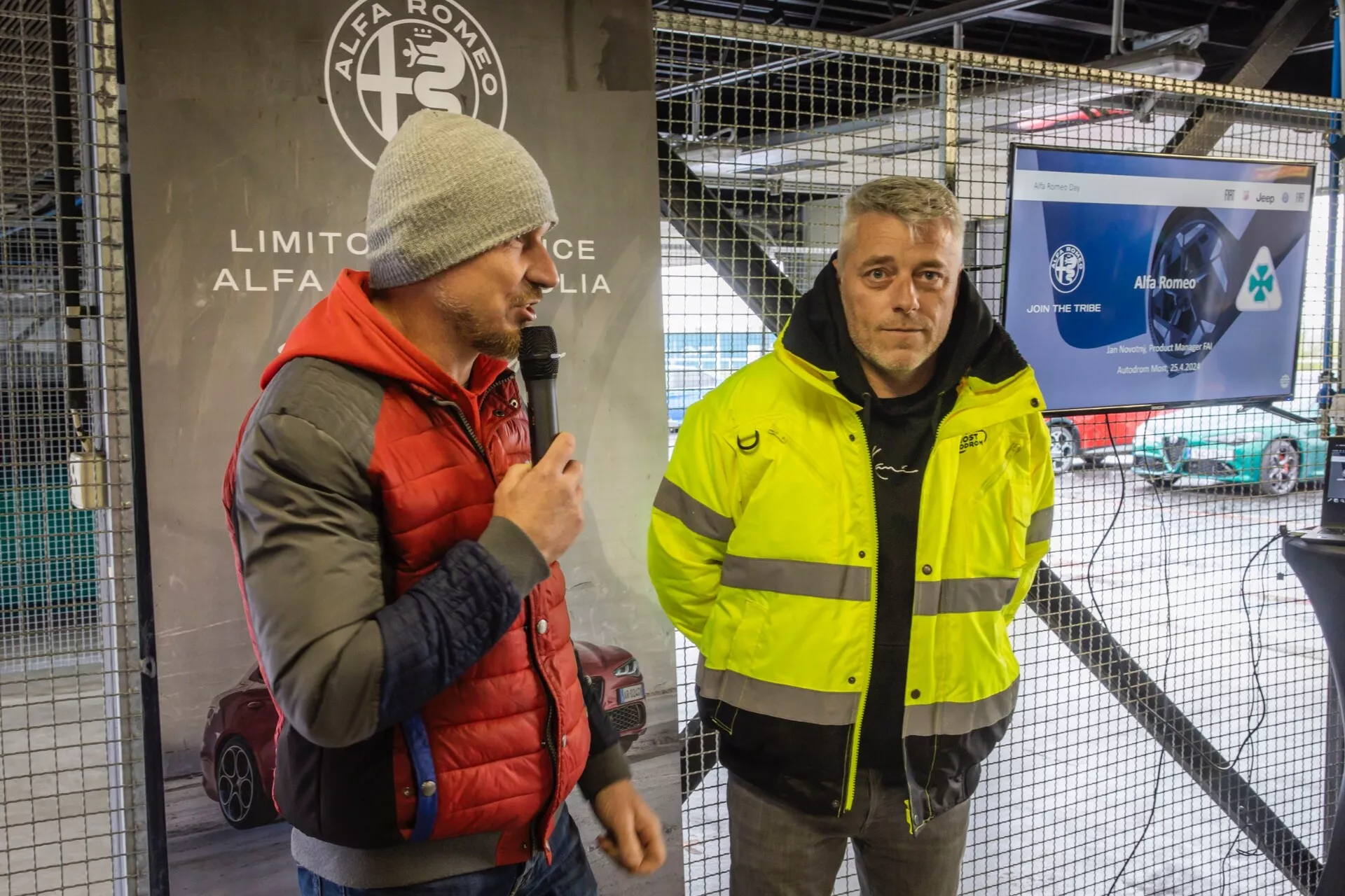 Deux hommes au premier plan, l'un parlant dans un microphone, l'autre écoutant attentivement, avec une voiture de course partiellement visible en arrière-plan, suggérant un événement de sport automobile