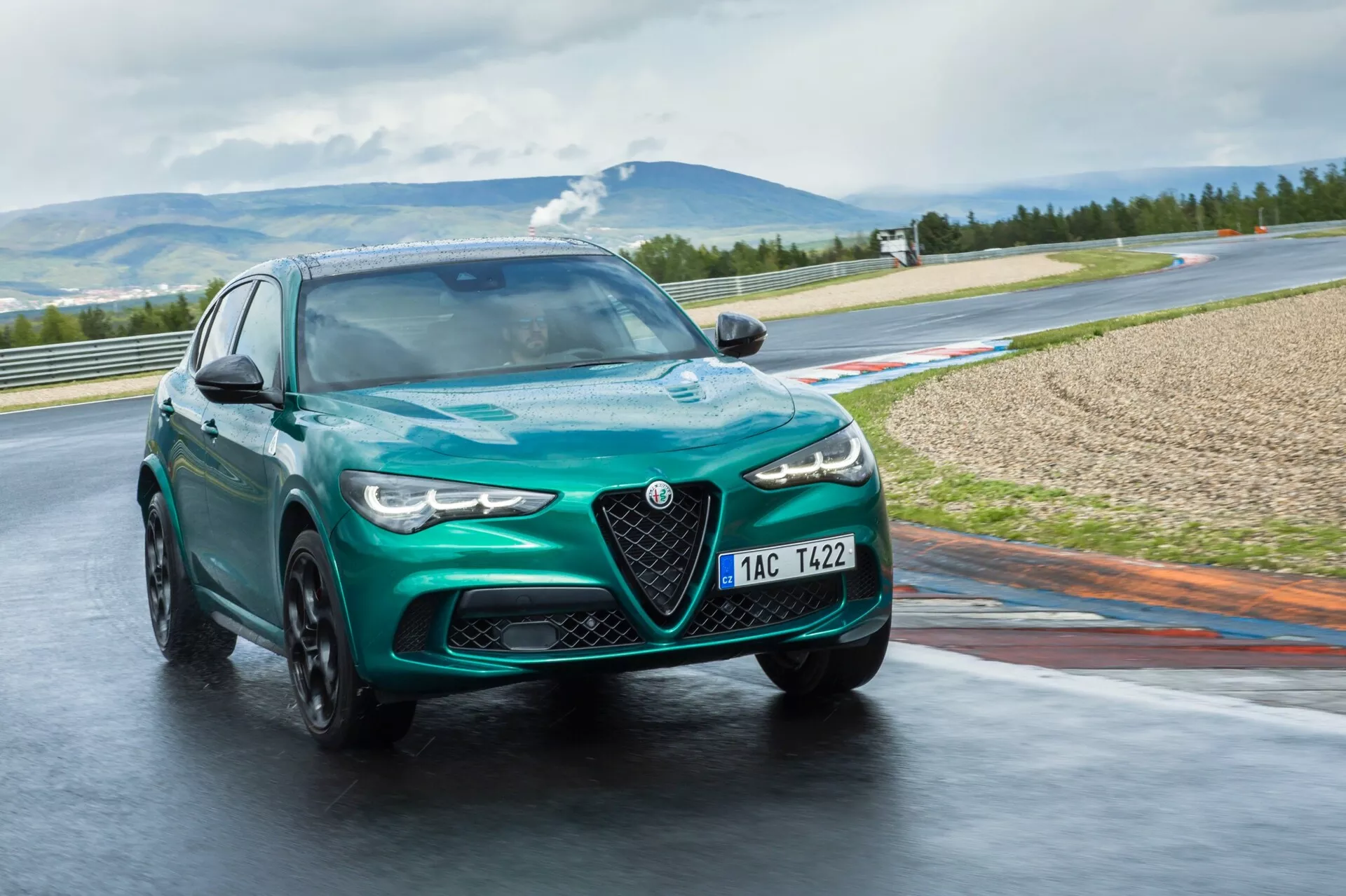Alfa Romeo Giulia Quadrifoglio 2023 en vert métallisé, capturée sous un angle dynamique de trois quarts avant sur une piste mouillée, mettant en avant les phares LED, les roues en alliage en mouvement et la grille Scudetto distinctive, sur un arrière-plan montagneux.