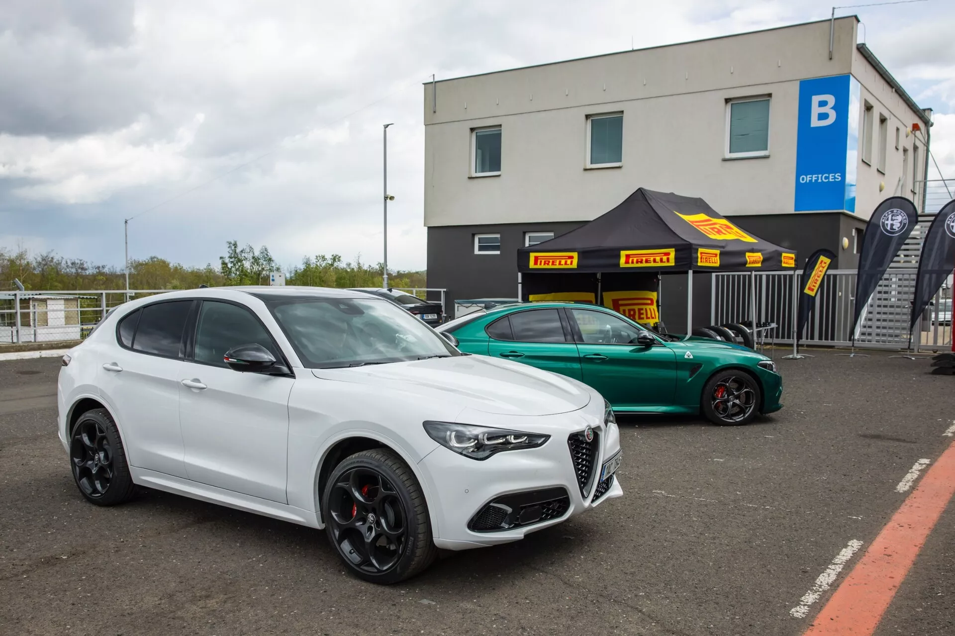 Alfa Romeo Giulia Quadrifoglio 2023 et SUV de luxe stationnés sur une surface en asphalte dans une zone potentiellement destinée à une piste de course ou un complexe industriel, affichant un badge distinctif de marque, des roues noires orientées performances et un style agressif sous un éclairage diurne nuageux