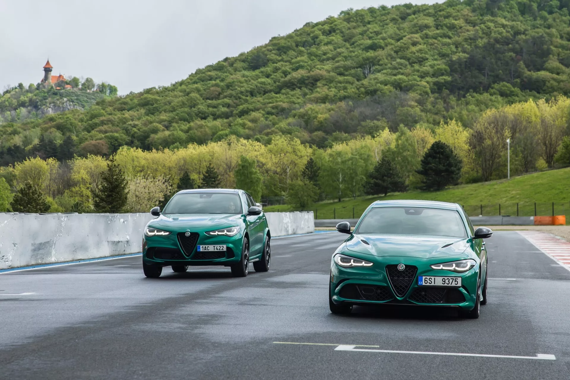 Vue dynamique de deux voitures Alfa-Romeo Giulia Quadrifoglio 2023 roulant sur piste, avec un focus détaillé sur la grille avant, les phares LED, et la conception aérodynamique contre un fond de verdure et de colline sous un ciel nuageux
