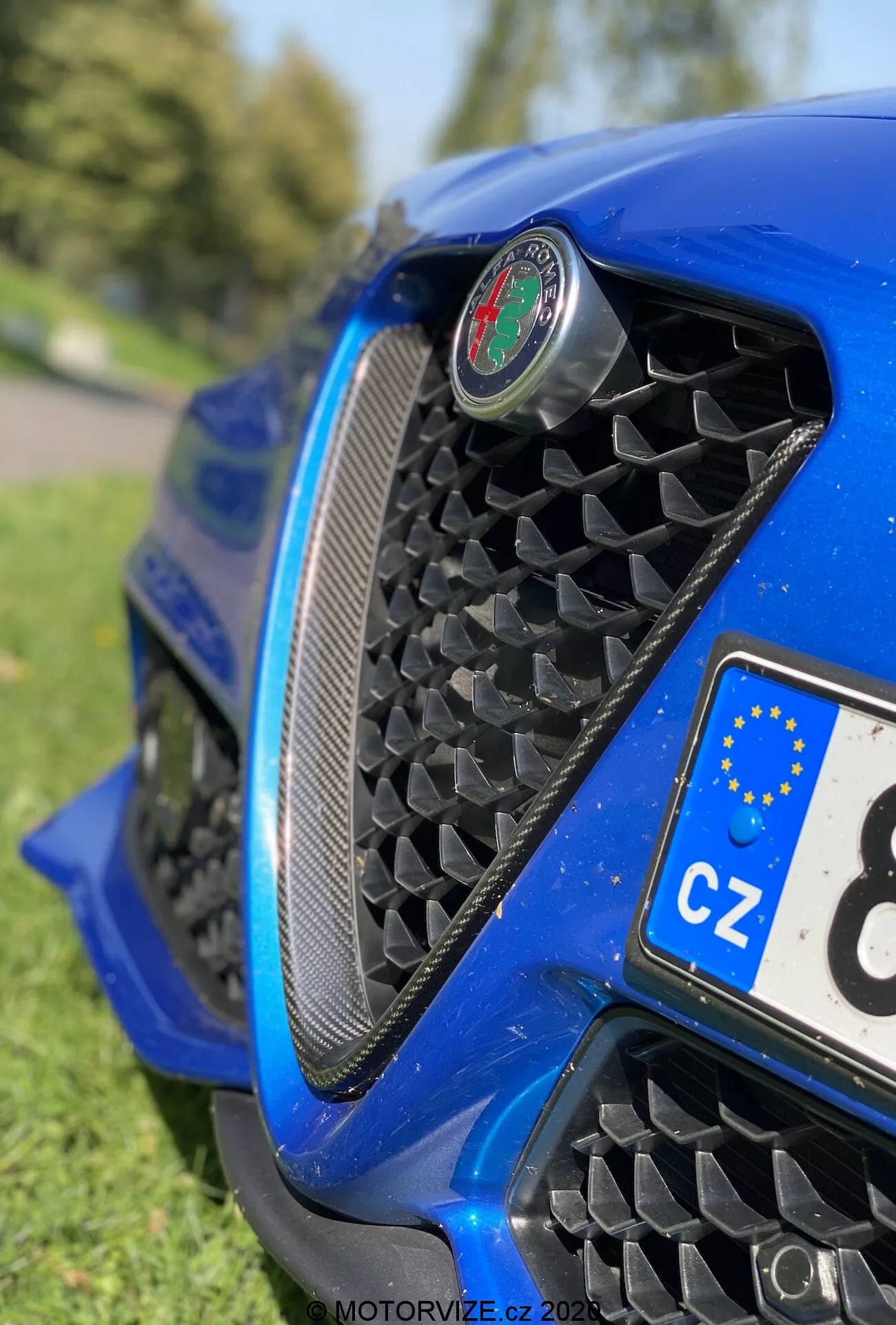 Vue rapprochée de l'avant de l'Alfa Romeo Giulia 2016 prise d'un angle bas, mettant en évidence la grille en nid d'abeille, l'emblème central, le pare-chocs avant aérodynamique et la carrosserie bleu métallisé brillant avec des reflets. Vue partielle d'une plaque d'immatriculation tchèque et arrière-plan flou rappelant un parc.