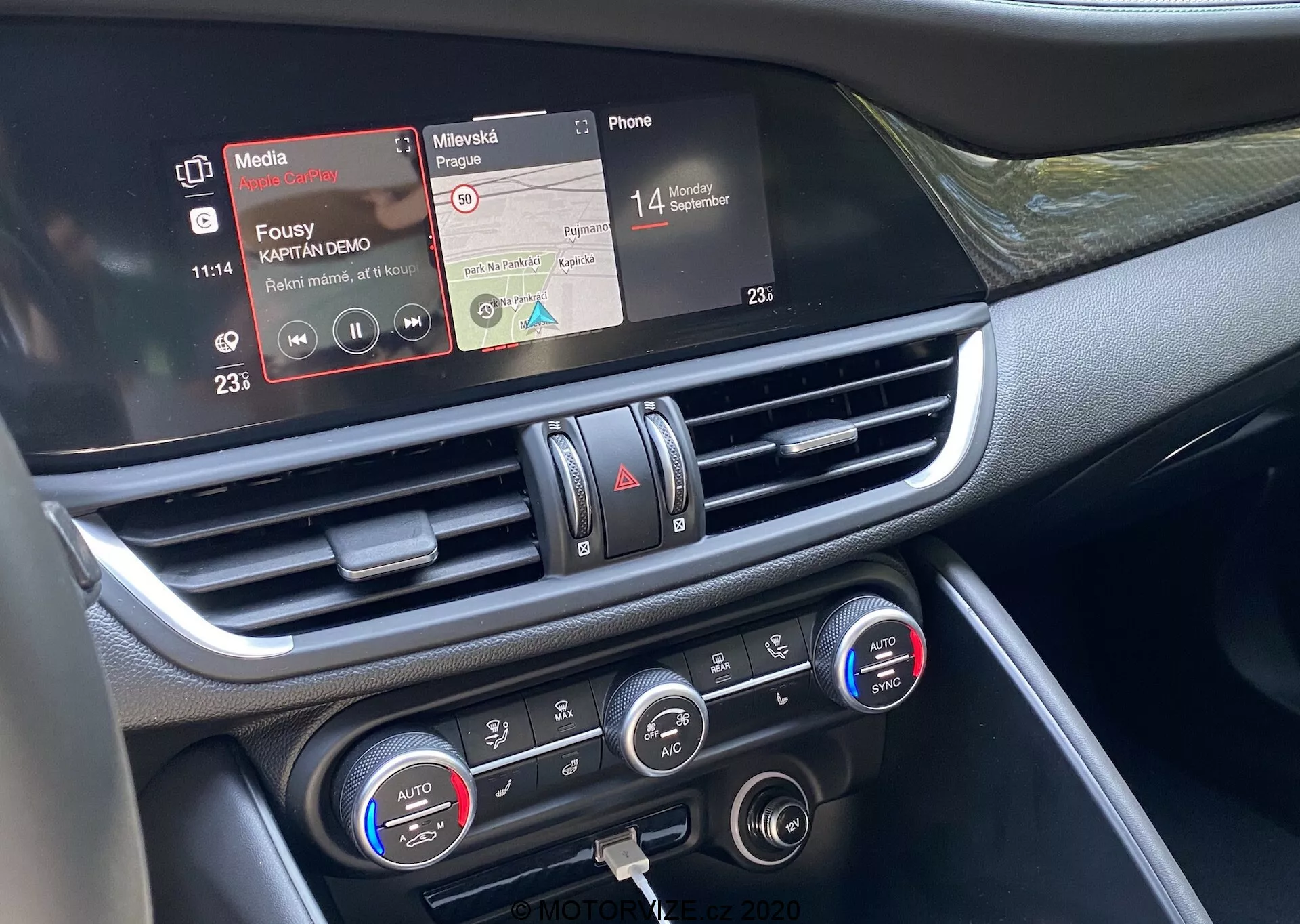 Vue rapprochée de la section centrale du tableau de bord de l'Alfa Romeo Giulia 2016 depuis le siège passager avant. L'image en gros plan met en évidence l'écran tactile d'infodivertissement avec les fonctionnalités de lecteur multimédia et de connectivité téléphonique, les bouches d'aération centrales avec des accents chromés et les commandes de climatisation avec des réglages de température à double zone et des affichages numériques. Les matériaux du tableau de bord semblent inclure des plastiques et des garnitures métalliques ou chromées, présentant la technologie intérieure avancée du véhicule axée sur la connectivité, la lecture multimédia et le contrôle de la climatisation.