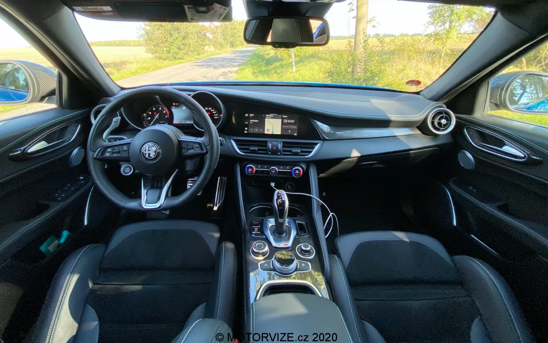 Vue intérieure d'une Alfa Romeo Giulia 2016 depuis la perspective du conducteur présentant un volant à fond plat avec plusieurs boutons de commande, un système d'infodivertissement numérique sur le tableau de bord, une horloge analogique, des aérateurs circulaires, un levier de vitesses élégant, un sélecteur de modes de conduite, des porte-gobelets, des vitres électriques, des sièges en cuir avec renforts latéraux et des caractéristiques technologiques modernes en général.