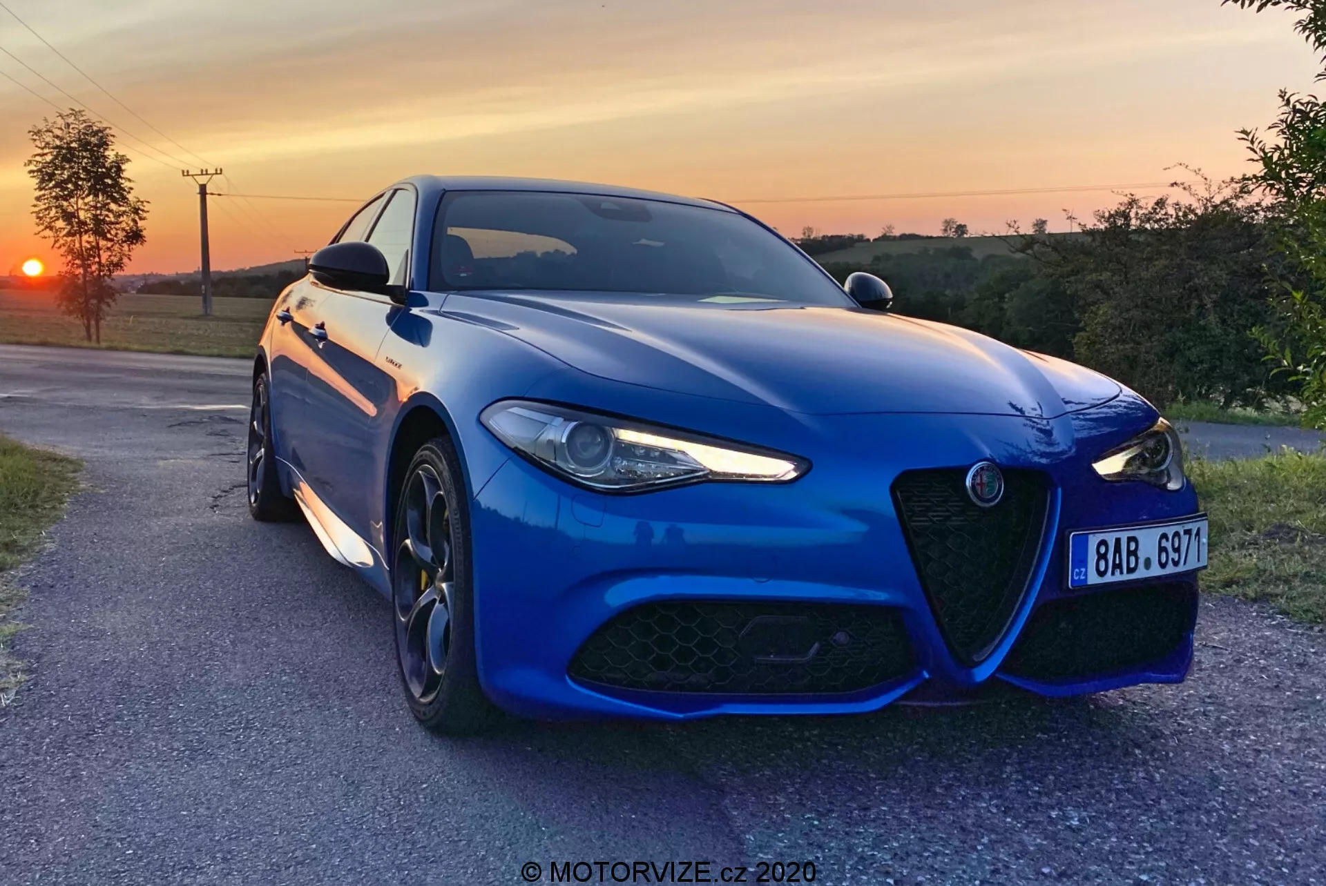 Une berline sportive Alfa-Romeo Giulia 2016 bleue capturée le soir avec un coucher de soleil en arrière-plan, positionnée légèrement en angle vers l'appareil photo sur une voie de campagne pavée, mettant en valeur sa calandre distinctive, son design agressif de phares à LED, ses grandes roues en alliage à plusieurs branches et son système de freinage haute performance. La peinture métallique, le pare-chocs avant aérodynamique, les lignes sculptées du capot et les capteurs avancés d'assistance à la conduite soulignent l'esthétique sportive et la technologie moderne de la voiture. Un filigrane 'MOTORVIZE.cz 2020' est présent.