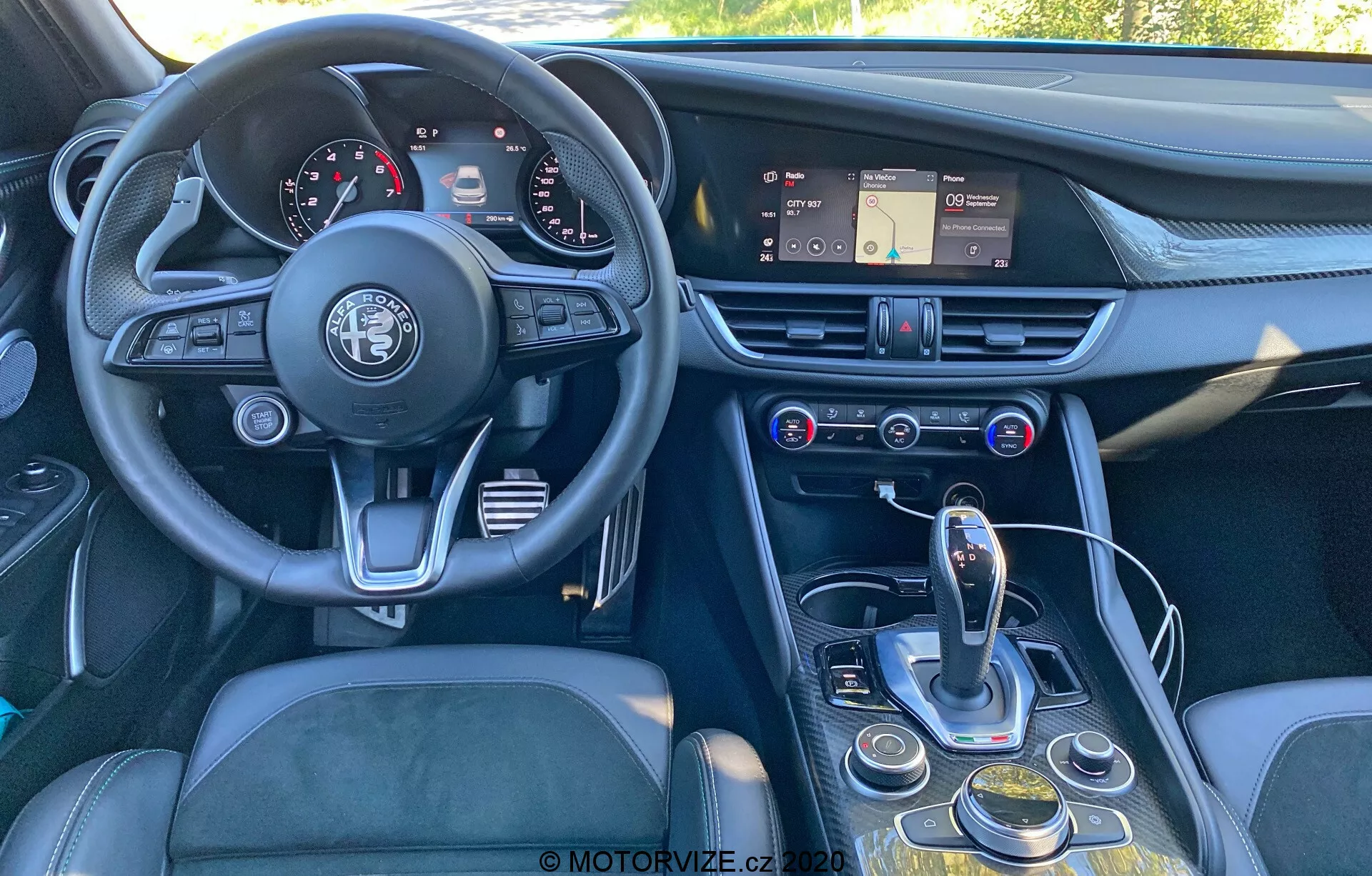 Vue intérieure d'une Alfa Romeo Giulia 2016 sous un angle légèrement décentré, de l'arrière vers l'avant, montrant le tableau de bord, le volant, la console centrale et une partie du siège conducteur. L'image met en évidence le volant à trois branches avec l'emblème Alfa Romeo, un tableau de bord avec des cadrans analogiques et des écrans numériques, un tableau de bord avec un système d'infodivertissement à écran tactile, des bouches d'aération et des commandes de climatisation et multimédia. La console centrale comprend un sélecteur de transmission automatique, un cadran rotatif, des boutons pour la navigation de l'infodivertissement et les réglages du véhicule, et un possible sélecteur de modes. Le design met l'accent sur l'ergonomie et l'accessibilité, avec des matériaux tels que le cuir, le métal brossé et le plastique de haute qualité pour une sensation de luxe.