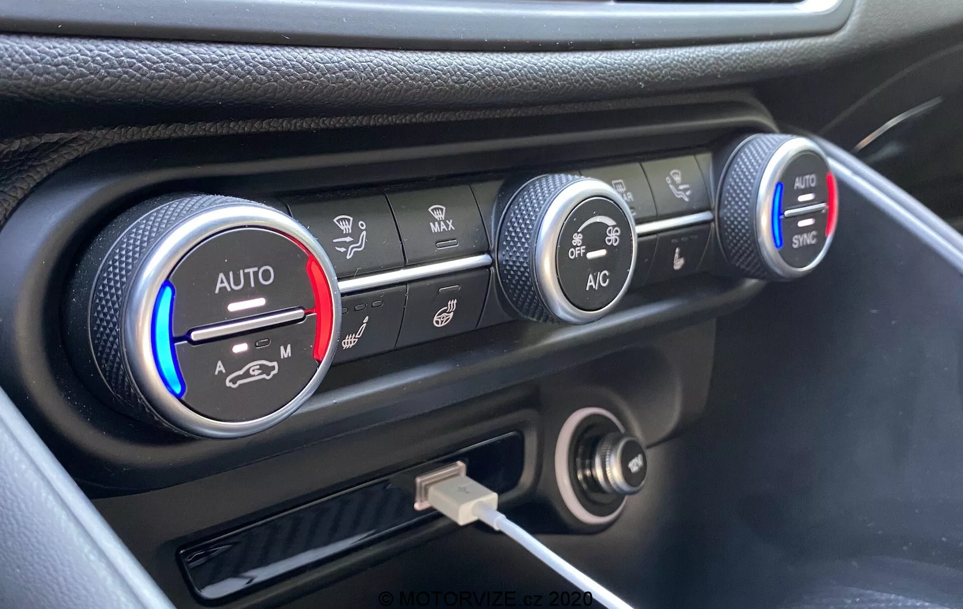 Gros plan du système de contrôle du climat sur le tableau de bord de l'Alfa Romeo Giulia 2016, avec trois boutons circulaires. Le bouton de gauche indique les réglages de température avec des bandes bleues et rouges et un réglage 'AUTO', partiellement éclairé en rouge. Le bouton du milieu affiche les réglages de la vitesse du ventilateur, mis en évidence à une vitesse inférieure. Le bouton droit montre les différents modes de distribution de l'air avec des boutons 'AC' et 'SYNC'. Sous les boutons se trouvent une prise de courant de 12 volts et un port USB. Le tableau de bord a une finition noire avec une touche argentée autour des commandes de climat, capturé sous un angle élevé.