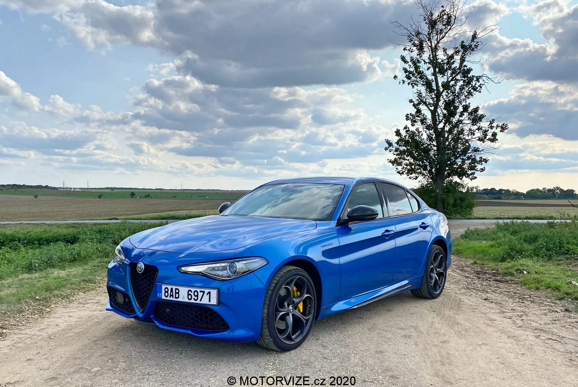 Image d'une berline sportive Alfa Romeo Giulia 2016 bleu vif garée sur un chemin de gravier, avec un ciel partiellement nuageux en arrière-plan. La voiture est capturée sous un angle avant trois-quarts, montrant sa calandre avant distinctive 'Trilobo', des étriers de frein jaunes, de grandes roues en alliage à rayons multiples, des phares à LED élégants et des entourages de fenêtres chromés. Le véhicule est légèrement décentré vers la gauche avec la roue avant gauche pointant vers l'extérieur, lui conférant une posture dynamique.