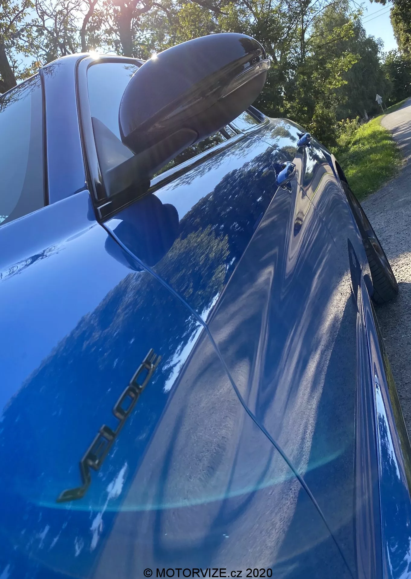 Image en gros plan d'une Alfa Romeo Giulia bleue (modèle 2016) photographiée sous un angle bas et vers le haut, se concentrant sur la section arrière. Les badges du modèle du côté conducteur, la peinture bleue réfléchissante, le feu arrière LED gauche, le becquet arrière noir, la vitre arrière inclinée avec le troisième feu stop, le reflet des arbres et du ciel lumineux et le rétroviseur avec clignotant intégré sont visibles.
