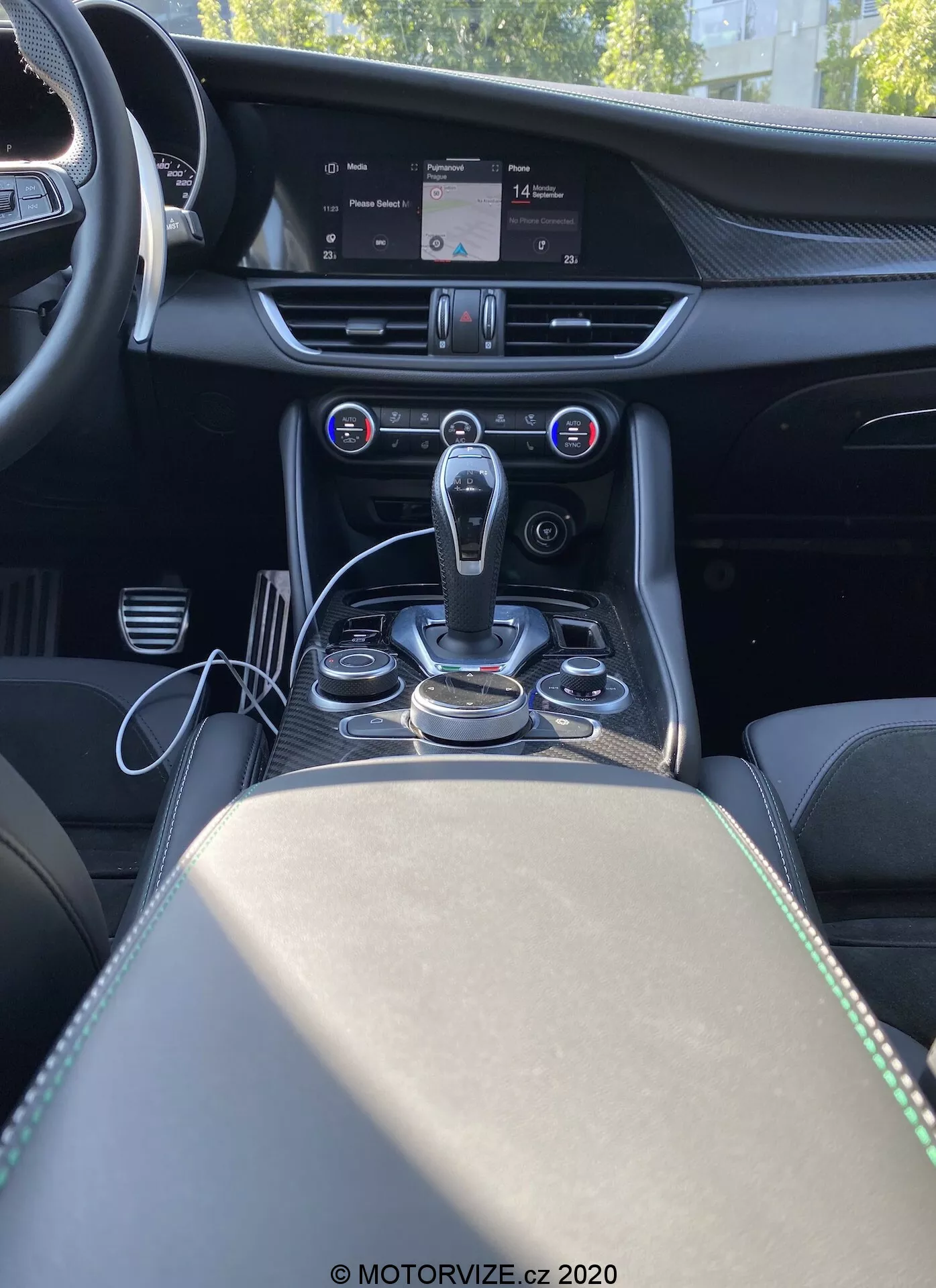 Capture d'intérieur de l'Alfa Romeo Giulia 2016 vue de derrière la console centrale, montrant la console centrale, le tunnel de transmission et une partie du siège passager. L'image offre une perspective frontale depuis le point de vue d'un passager arrière, mettant en évidence un levier de vitesses automatique moderne entouré de cadrans et de boutons de commande métalliques pour le système d'infodivertissement et les modes de conduite. Au-dessus de l'espace de changement de vitesses se trouve le système de contrôle climatique automatique à deux zones avec un écran numérique affichant les réglages de température et des boutons pour contrôler la distribution de l'air et la vitesse du ventilateur. Un grand écran d'infodivertissement au sommet de la console centrale affiche une carte du système de navigation. Le tableau de bord présente des détails de couture et des accents métalliques qui suggèrent des matériaux haut de gamme. L'accoudoir et le compartiment de rangement avec deux porte-gobelets sont visibles derrière le levier de vitesse, enrichissant le design luxueux et axé sur la technologie de l'habitacle.