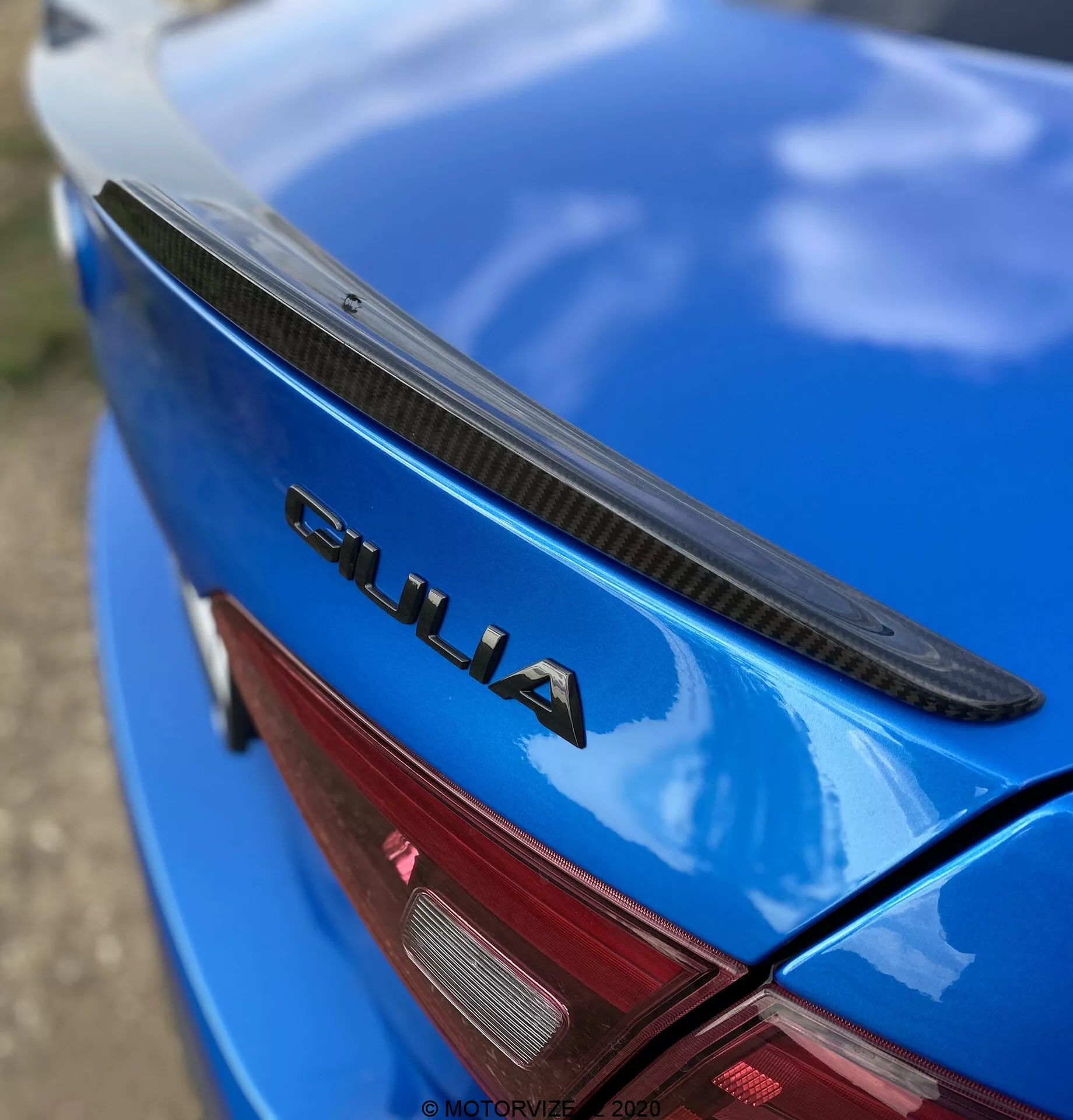 Vue en gros plan et en angle de la section arrière d'une Alfa Romeo Giulia 2016 bleue. La photographie montre le couvercle du coffre avec un spoiler noir, une peinture bleue brillante et un badge chromé 'GIULIA' bien en évidence. Une partie du feu arrière LED rouge est également visible. Le badge 'GIULIA' est nettement focalisé, tandis que le spoiler, la surface de la voiture et le feu arrière s'estompent graduellement, mettant en valeur les éléments de la marque et du design de la voiture.