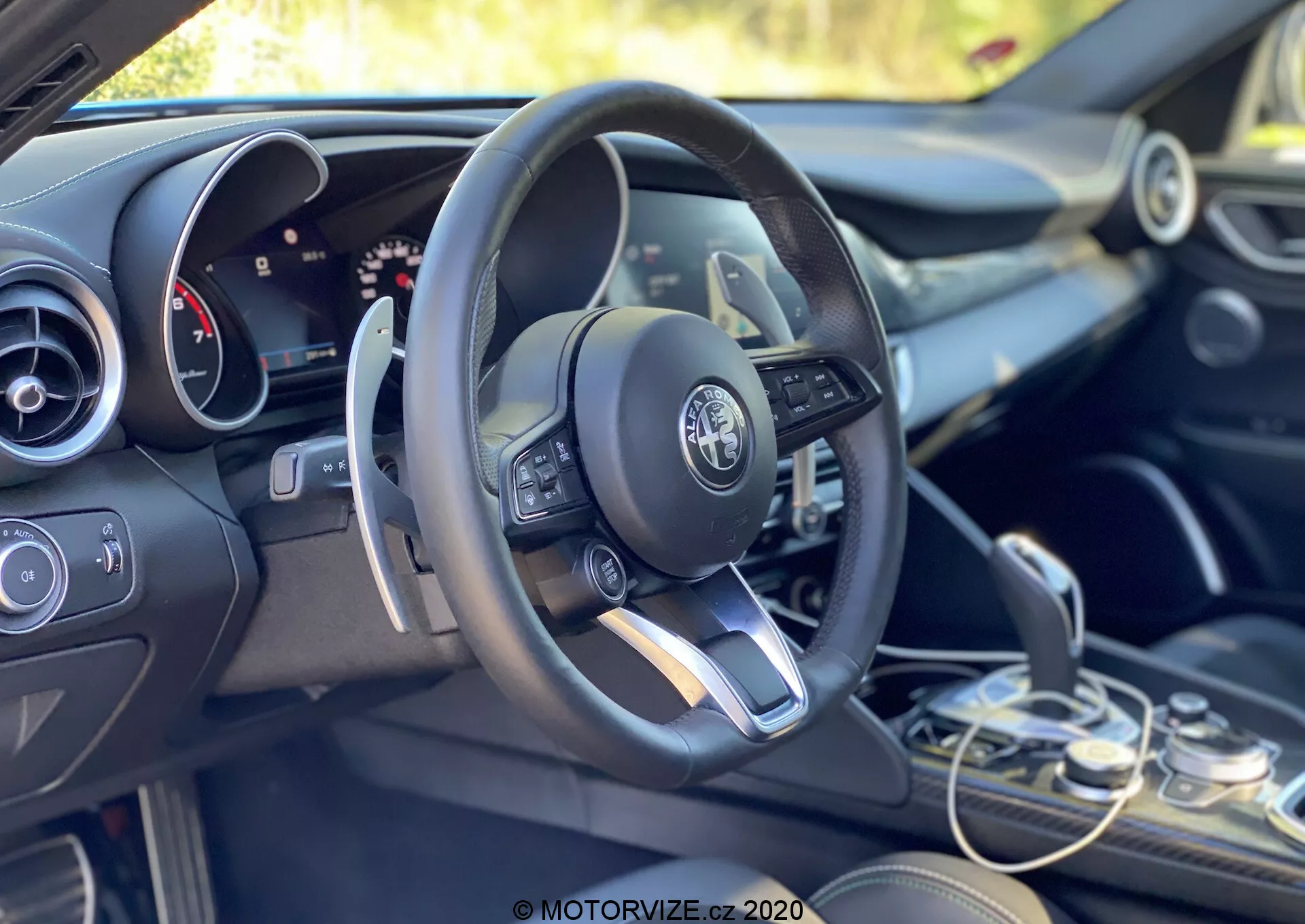 Vue intérieure d'une Alfa Romeo Giulia 2016 centrée sur la zone du conducteur et le tableau de bord depuis une perspective inclinée vers l'arrière, côté gauche. La configuration de conduite à gauche présente un volant multifonction moderne avec des commandes intégrées, incluant des boutons pour régler le volume, accéder aux menus et probablement le régulateur de vitesse. Le logo Alfa Romeo est clairement visible au centre du volant. Derrière le volant, partiellement visible, se trouve un tableau de bord mixte avec affichages analogiques et numériques, indiquant un système d'information configurable pour le conducteur. À droite du volant, la partie supérieure de l'écran du système d'infodivertissement est à peine visible, flanquée par les contrôles du système de climatisation et les interrupteurs fonctionnels en dessous. Le sélecteur de vitesse est situé dans le coin inférieur droit, affichant un design élégant de transmission automatique. Les finitions de l'intérieur combinent des tons sombres avec des accents métalliques, reflétant le design premium du véhicule. L'habitacle est baigné de lumière naturelle, avec un arrière-plan vert suggérant un environnement extérieur ou végétatif.