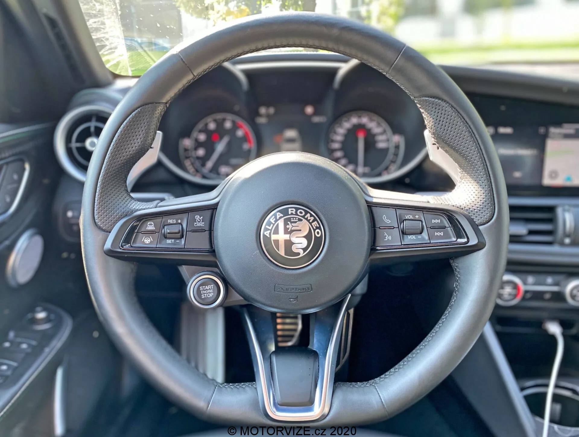 Perspective du conducteur de l'intérieur de l'Alfa-Romeo Giulia 2016, se concentrant sur le volant et le tableau de bord. Le volant multifonction centrallement positionné dispose de boutons pour les médias, le contrôle vocal, la connectivité téléphonique, les informations sur le véhicule et les réglages du régulateur de vitesse. Le tableau de bord affiche des cadrans analogiques, probablement un compteur de vitesse et un compte-tours, avec un écran numérique entre les deux. Le volant combine cuir, coutures et accents métalliques, évoquant le luxe. Les éléments en arrière-plan comprennent les commandes du système de climatisation et un écran central au-dessus.
