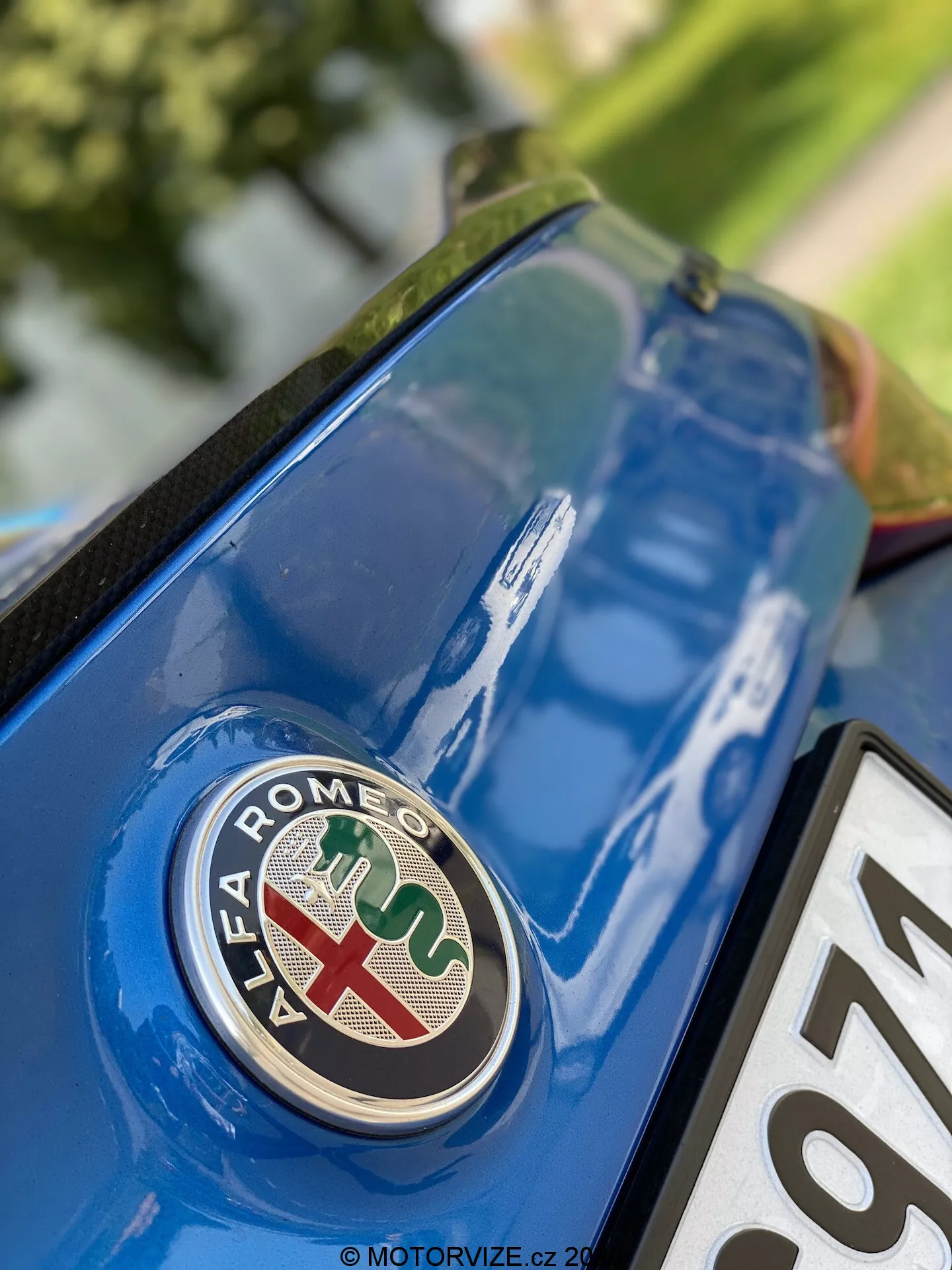 Vue rapprochée de la section arrière de l'Alfa Romeo Giulia 2016 avec un focus sur l'emblème de la marque depuis un angle bas. Le badge, avec une croix rouge sur fond blanc et un serpent vert sur fond bleu, est net sur le couvercle de coffre bleu métallisé, avec des détails de surface environnants et des reflets.
