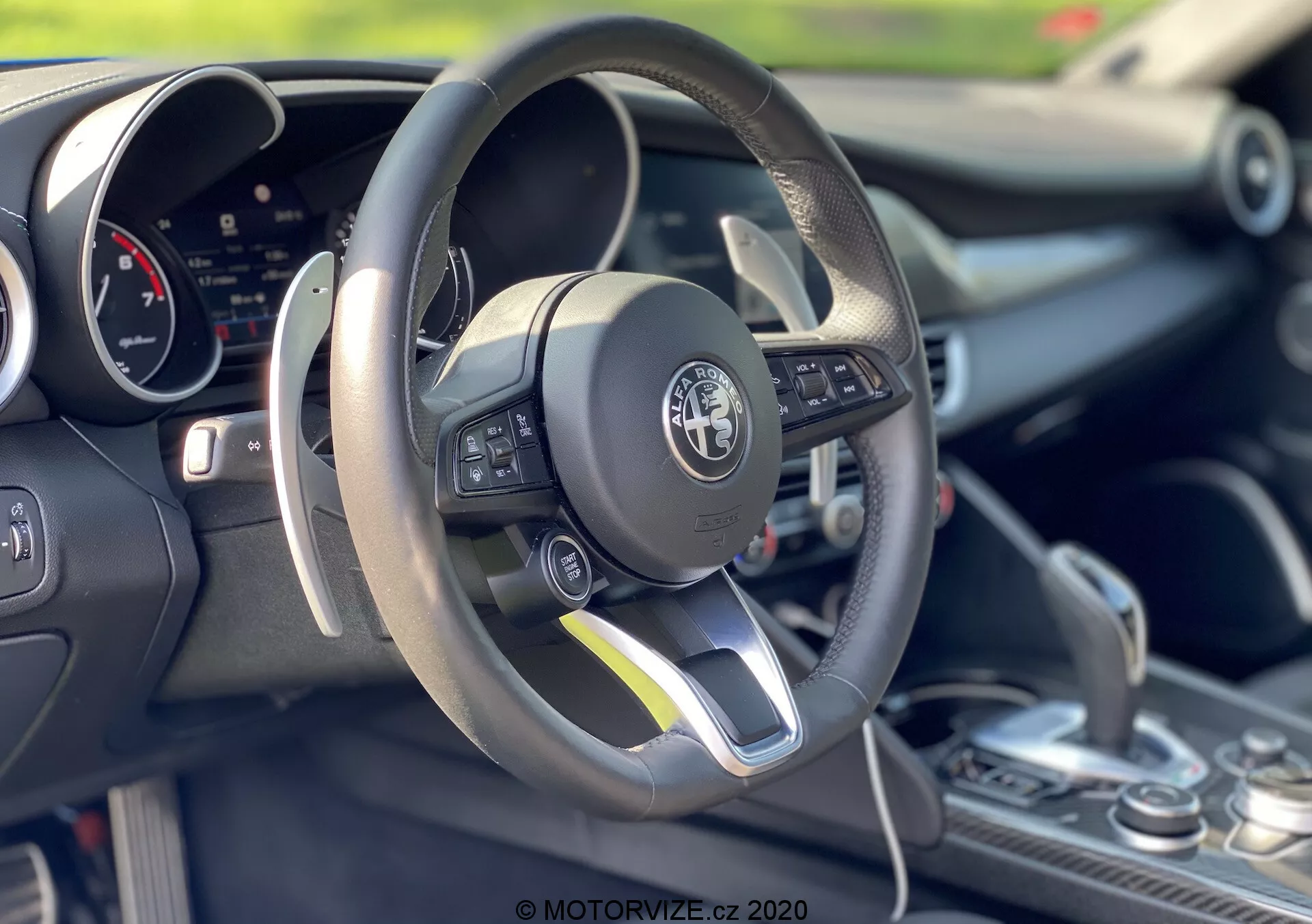 Photo intérieure d'une Alfa-Romeo Giulia 2016 se concentrant sur la zone du conducteur et le volant. Le volant à fond plat, recouvert de cuir, comporte des commandes multifonctions sur le côté gauche. Derrière le volant, on aperçoit un tableau de bord numérique. La console centrale montre un levier de vitesses avec un soufflet en cuir, suggérant d'éventuelles capacités de changement manuel. L'image met en avant le design sportif de la voiture, la technologie avancée et la minutie de fabrication avec une faible profondeur de champ mettant en valeur les éléments du premier plan.