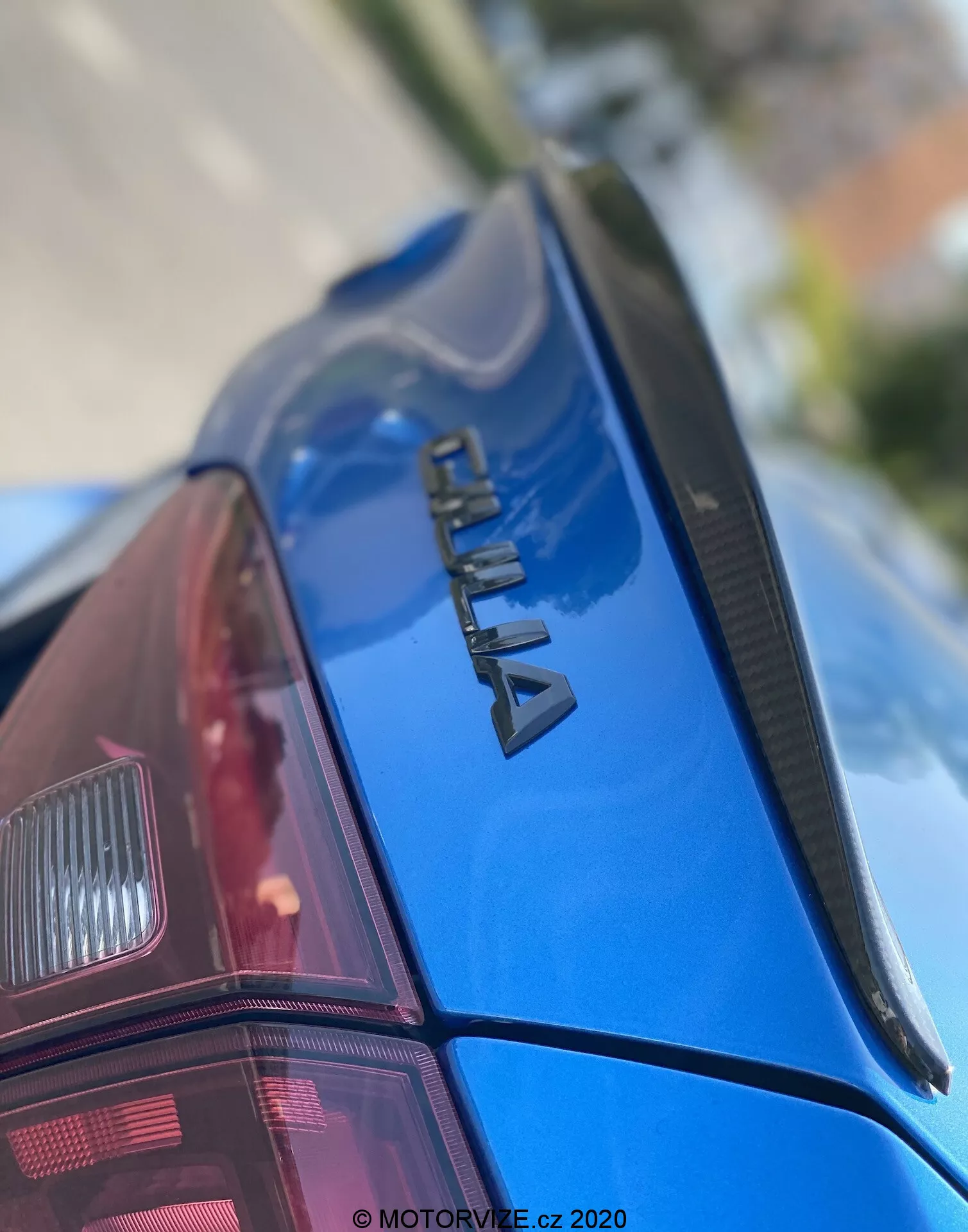 Gros plan de la section arrière d'une voiture bleue ŠKODA, vue d'un angle vers le haut. L'accent est mis sur le logo chromé 'ŠKODA' sur le couvercle du coffre, un spoiler fin et la peinture brillante et bien entretenue. Le pare-brise arrière présente des lignes de dégivrage et partiellement visible le groupe de feux arrière moderne. La faible profondeur de champ met en évidence la marque, tandis que l'avant et l'arrière-plan sont flous. Filigrane 'MOTORVIZE.cz 2020', indiquant la date de publication ou de capture.