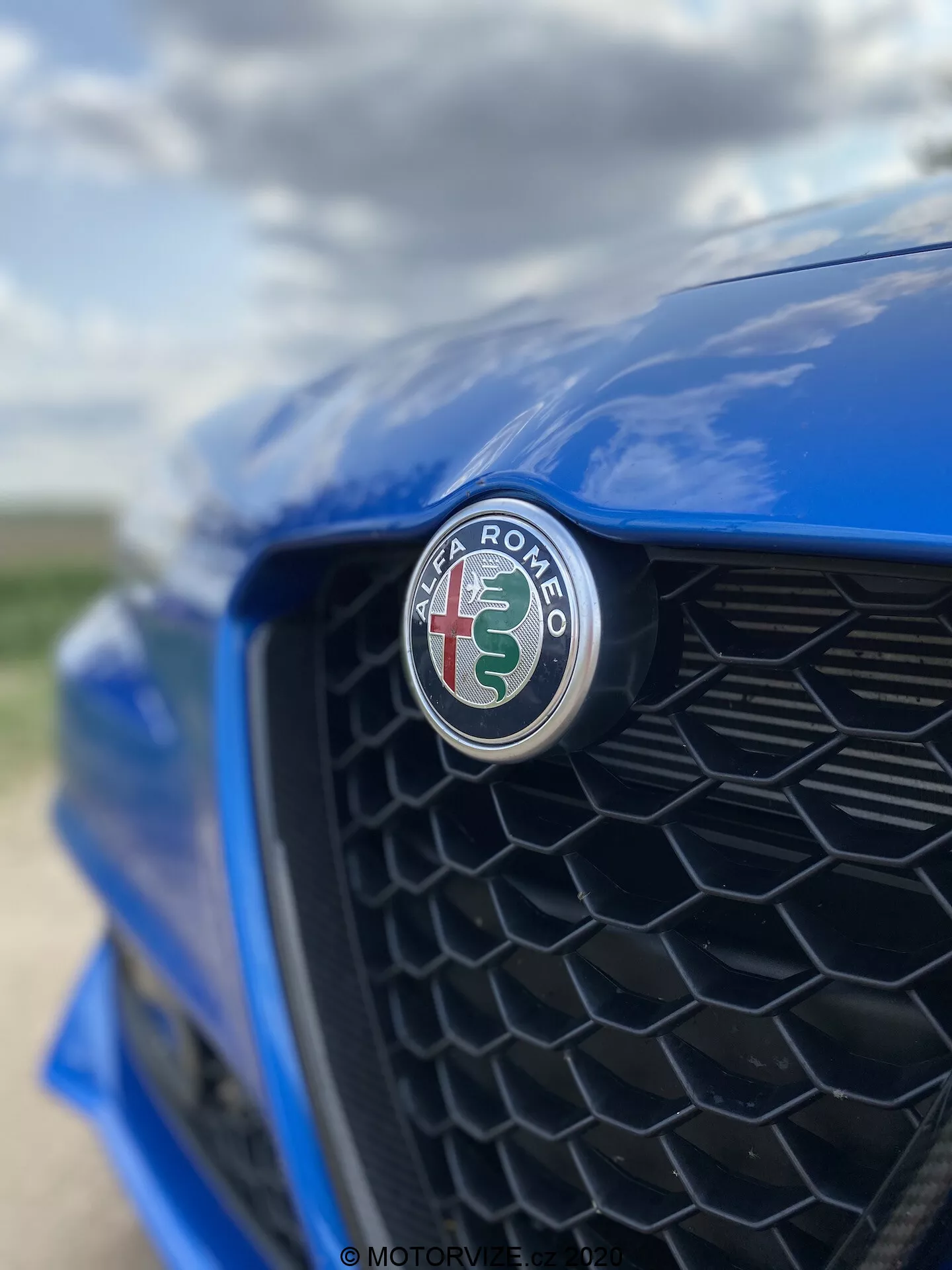 Gros plan de la section avant d'une Alfa Romeo Giulia 2016 bleue, se concentrant sur l'emblème du fabricant avec une croix rouge et un serpent vert sur un fond blanc. Pris sous un angle décentré vers le côté conducteur, la grille noire en nid d'abeille est légèrement floue, soulignant la netteté de l'emblème et la finition brillante.