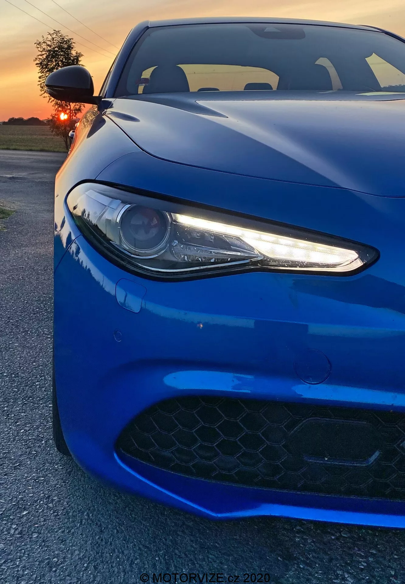 L'image montre une voiture de sport Alfa Romeo Giulia 2016 capturée sous un angle frontal bas. Le véhicule bleu profond vibrant remplit le cadre, mettant en avant son phare gauche avec les feux de jour LED, une partie du capot et la grille d'admission d'air du pare-chocs avant. Le phare de type projecteur, avec ses accents LED blancs élancés, donne un aspect agressif et moderne. La grille noire à motif hexagonal suggère des capacités de haute performance. La prise de vue capture les lignes aérodynamiques et les contours de la voiture, avec des reflets du coucher de soleil accentuant sa finition brillante. En arrière-plan, on voit un ciel orange éclairé partiellement occulté par le soleil. L'angle rapproché du photographe met en avant les détails de conception, évoquant un sentiment de puissance et d'élégance.