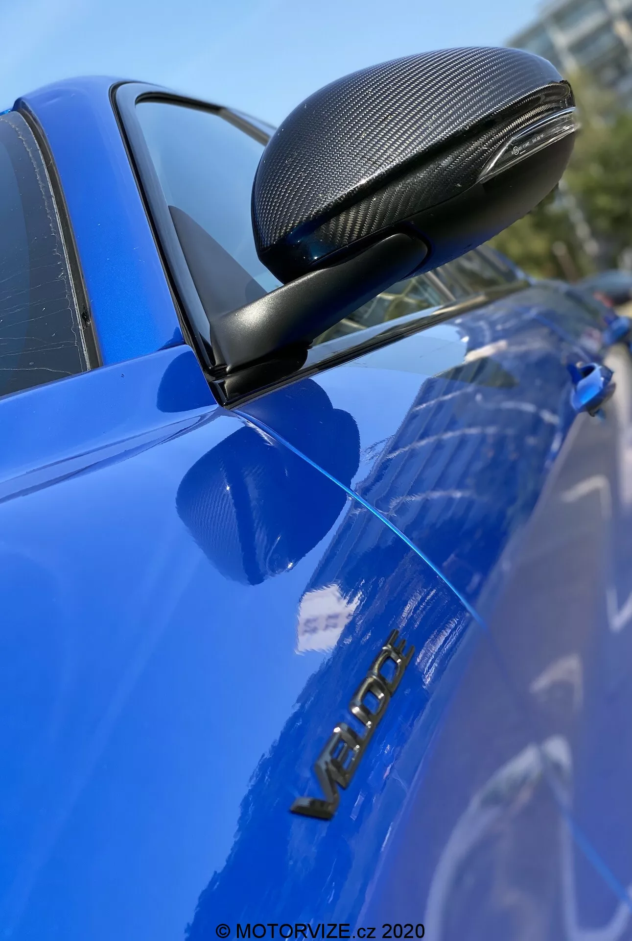 Image en gros plan d'une Alfa Romeo Giulia 2016 bleue, mettant en valeur le rétroviseur latéral avec une finition en fibre de carbone dans un carénage aérodynamique, monté sur un support noir. La photo se concentre nettement sur le rétroviseur, la porte du conducteur et le badge métallique 'VERON', soulignant ces détails avec une faible profondeur de champ et une surface brillante et bien entretenue.