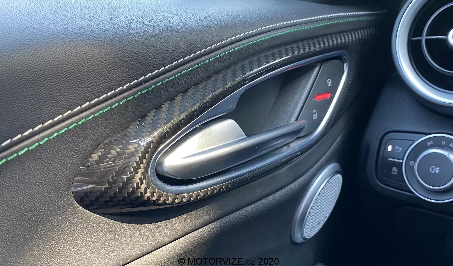Vue intérieure de la porte côté conducteur de l'Alfa Romeo Giulia 2016 depuis la perspective de quelqu'un assis à l'intérieur, mettant en avant une poignée de porte métallique placée dans un renfoncement à motif en fibre de carbone, des coutures vertes sur le panneau de porte en cuir, des boutons de contrôle pour les rétroviseurs latéraux et les fenêtres, une grille d'enceinte et des finitions en cuir et fibre de carbone de haute qualité indiquant un véhicule de luxe ou de performance.
