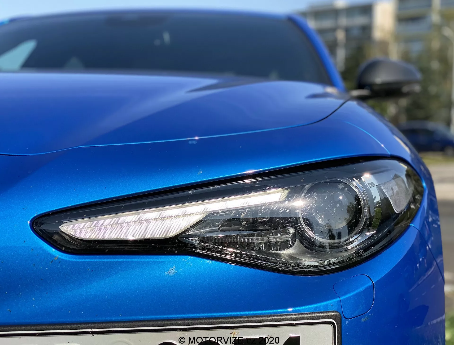Gros plan sur le côté avant gauche d'une Alfa Romeo Giulia 2016 bleue, capturé d'un angle bas mettant en évidence les détails du phare et la courbure de la voiture. Le phare moderne présente un éclairage LED ou HID avec une forme angulaire élégante et un faisceau principal de style projecteur pour une lumière focalisée. Le capot a un design lisse et profilé avec une finition métallique réfléchissante, et le pare-chocs avant exhibe un design minimaliste. Le rétroviseur latéral, de la même couleur que la carrosserie et profilé, est partiellement visible dans le coin supérieur droit, complétant le profil aérodynamique de la voiture.