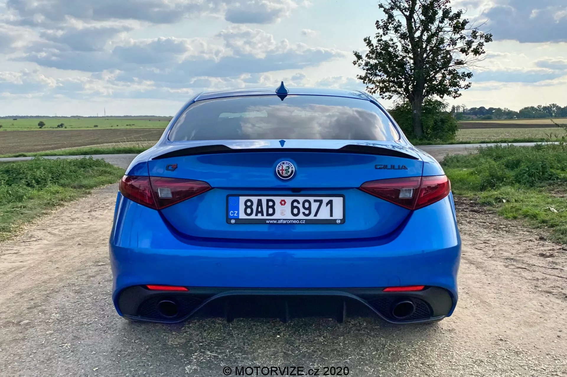 Vue arrière d'une berline Alfa Romeo Giulia 2016 bleu vif, prise d'une perspective au niveau des yeux. L'image montre tout l'arrière de la voiture, centrée dans le cadre, avec la plaque d'immatriculation, les feux arrière et le logo du fabricant clairement visibles. Les caractéristiques incluent un système d'échappement double, des feux arrière modernes et élégants, un élément diffuseur sur le pare-chocs, un spoiler sur le couvercle du coffre, des capteurs de stationnement et un essuie-glace arrière, ce qui suggère un équilibre entre caractère sportif, sécurité et praticité. Le décor est une zone rurale avec un paysage plat et un ciel partiellement nuageux pendant la journée.
