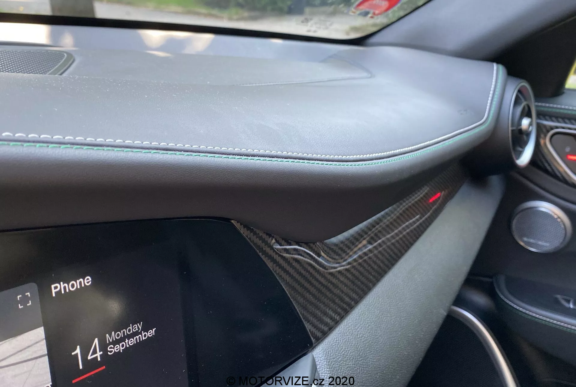 Gros plan de l'intérieur de l'Alfa Romeo Giulia 2016 depuis la perspective du passager, se concentrant sur le tableau de bord avec un matériau semblable au cuir et des coutures visibles, une garniture semblable à la fibre de carbone, un écran d'info-divertissement partiellement visible affichant des options de connectivité téléphonique avec le texte 'Phone' et 'Monday 14 September'. Voyant rouge au-dessus du système d'info-divertissement indiquant un système de sécurité actif, mettant l'accent sur le luxe et le design moderne.