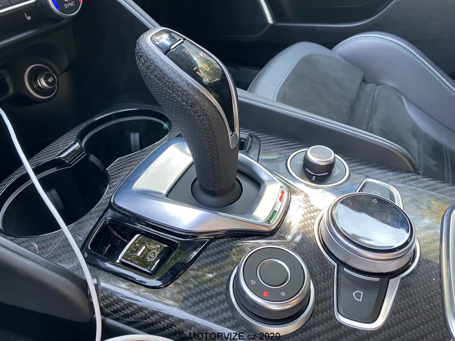 Vue rapprochée de la console centrale et du levier de vitesse de l'Alfa Romeo Giulia 2016, avec un levier de vitesse automatique gainé de cuir avec des accents chromés, entouré de finitions en fibre de carbone. La console comprend un bouton de contrôle de type iDrive, un bouton de volume, des boutons de dynamique de conduite, un port USB avec un câble connecté et un panneau avec les commandes des feux de détresse et autres, mettant en avant le design ergonomique et technologique de l'intérieur de la voiture.