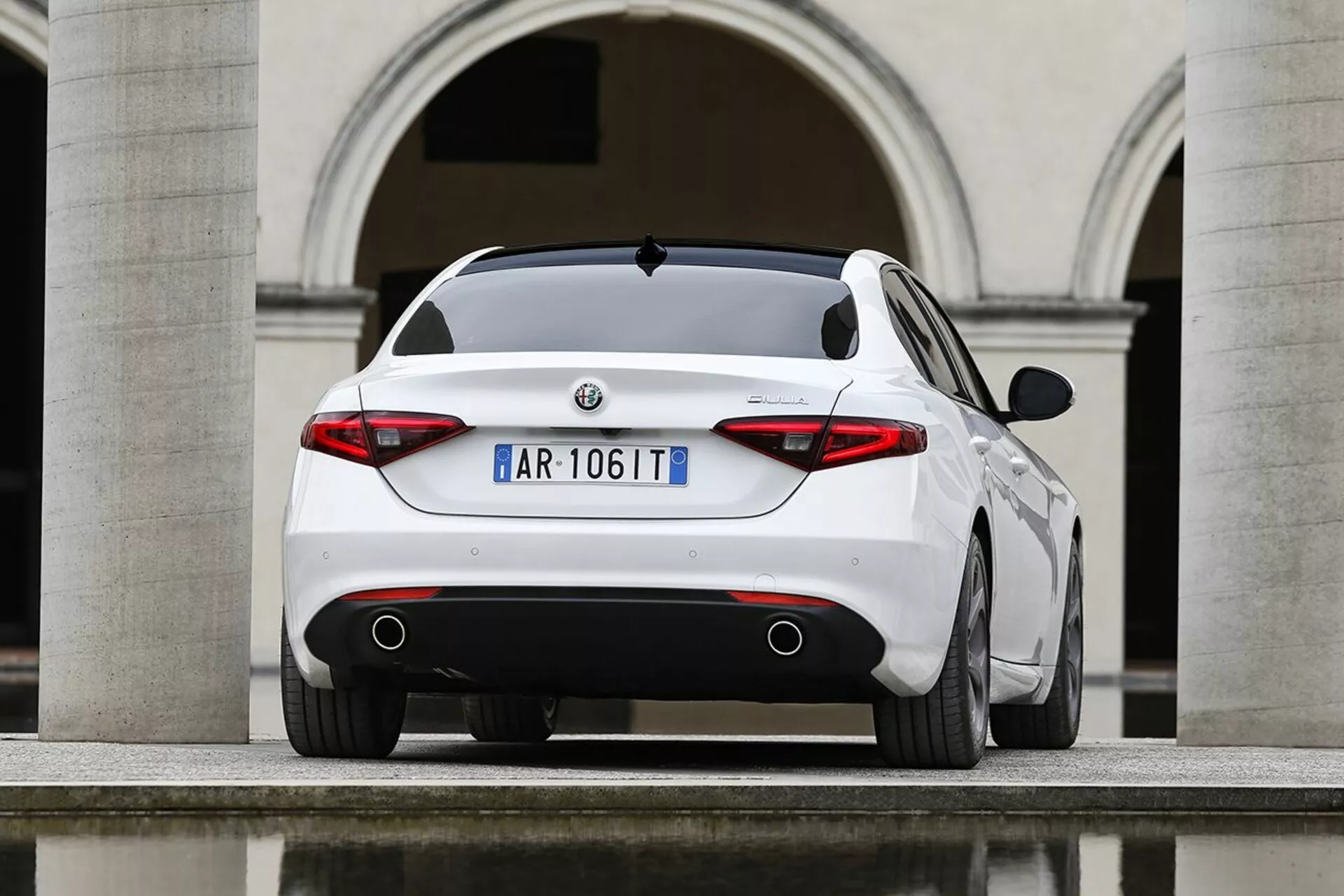 Alfa Romeo Giulia blanche de 2016 photographiée sous un angle arrière trois quarts mettant en évidence son profil arrière sportif, ses feux arrière distinctifs, ses doubles tuyaux d'échappement et ses éléments de design élégants sur fond d'architecture européenne