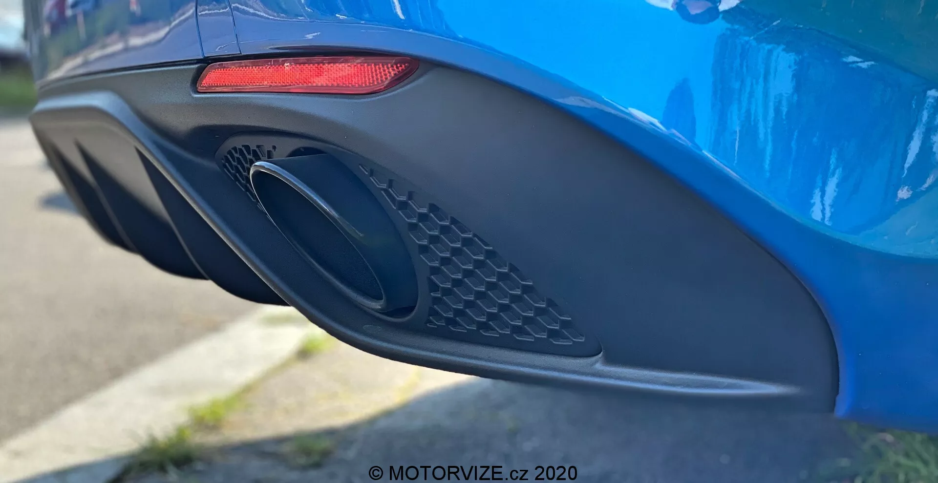 Vue latérale en gros plan de la zone arrière du pare-chocs et de l’échappement d'une Alfa Romeo Giulia 2016 bleu vibrant, mettant en valeur ses caractéristiques de design moderne, y compris un feu arrière LED horizontal rouge allongé, une grande sortie d'échappement ovale avec un détail de grille, et un pare-chocs aérodynamique avec diverses courbes et indentations, photographié avec une faible profondeur de champ pour accentuer les détails de la voiture.