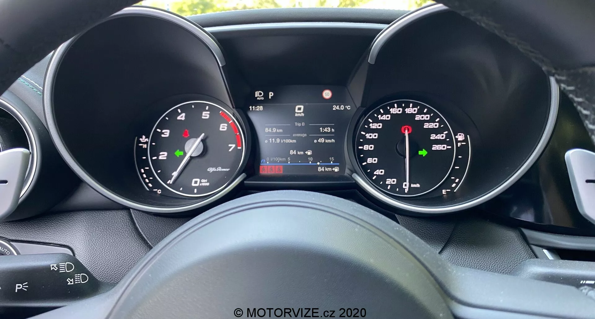 Vue du conducteur sur le tableau de bord de l'Alfa Romeo Giulia 2016, avec un écran central numérique affichant la vitesse, le rapport, la température et le kilométrage, encadré par des jauges analogiques de vitesse et de régime, présentant un mélange de design traditionnel et de technologie moderne, avec des vues partielles des commandes au volant pour l'audio et le régulateur de vitesse.