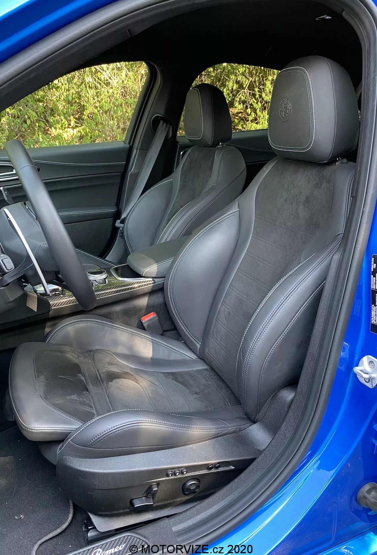 Vue détaillée du siège conducteur sportif dans une Alfa-Romeo Giulia 2016, montrant les réglages manuels du siège, les garnitures en cuir et en suédine, le logo embossé sur l'appuie-tête, une légère usure, le volant partiellement visible recouvert de cuir et les garnitures de porte assorties, pris sous un angle extérieur au-dessus du niveau du siège avec un intérieur sombre et un extérieur bleu vif contrastant.